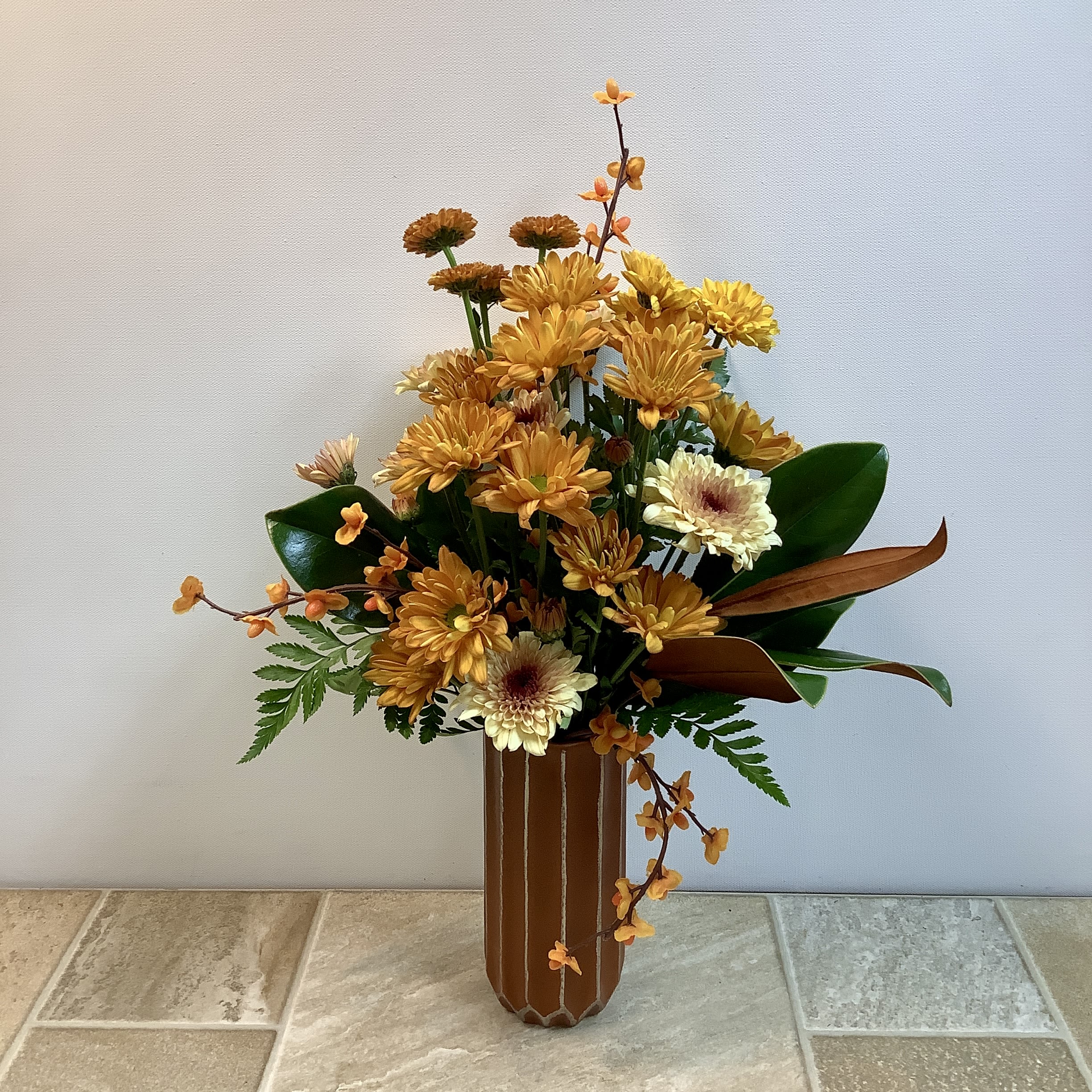Golden Sunset Golden Sunset - This arrangement has a warm, autumn-inspired look with a mix of golden, orange, and creamy tones. It features a variety of chrysanthemums in shades of amber, yellow, and pale cream, accented with small orange blooms and textural greenery. The glossy magnolia leaves add depth and richness, with their green tops and velvety brown undersides, while fern foliage softens the edges. The tall, fluted terracotta-colored vase with subtle striping complements the earthy color palette beautifully, giving the design a rustic yet elegant feel. It measures approximately 14” wide x 20” tall.