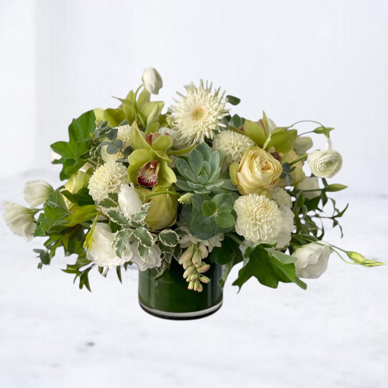 Verdant Serenity - The Verdant Serenity arrangement radiates tranquility and sophistication, combining soft greens and whites for a harmonious design. Featuring exotic green cymbidium orchids, creamy white ranunculus, lush hydrangeas, and white lisianthus, this bouquet is accented with textured greenery for a contemporary and refined look.  Perfect for milestones, celebrations, or to bring a touch of serenity to any setting, this elegant floral design embodies understated luxury. Expertly handcrafted, this arrangement makes a thoughtful gift for birthdays, anniversaries, or just to brighten someone’s day with the timeless beauty of nature.