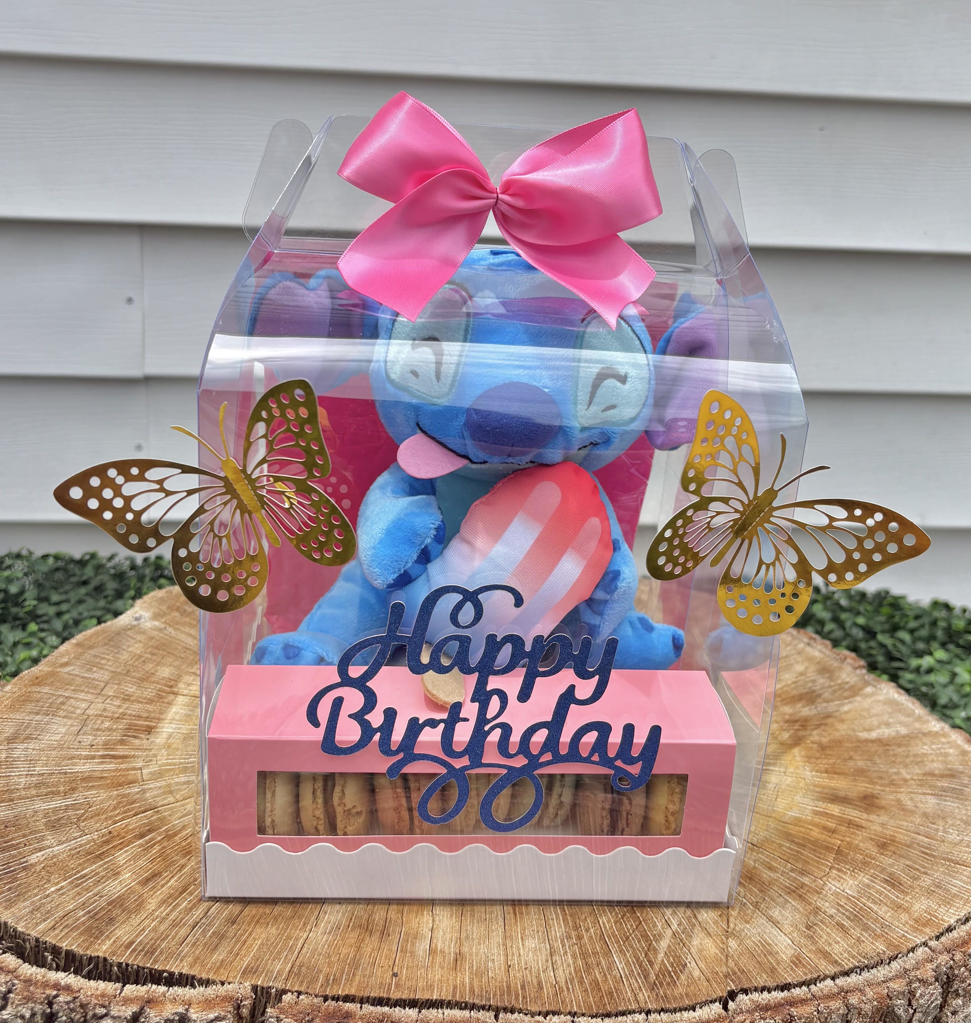 Sweet Stitch Surprise - A clear gift box featuring an adorable blue Stitch plushie paired with a delightful macaron set is more than just a present—it’s a joyful experience. Here’s why it might be the perfect choice for someone celebrating a loved one’s birthday:  Emotionally Uplifting  The Stitch plush brings instant smiles—it’s cute, comforting, and evokes fond memories for fans of the character  It offers a sense of nostalgia and whimsy, especially for someone who loves playful or animated gifts  Birthday Celebration in a Box  The Happy Birthday banner adds festive flair, making it feel like a party in miniature  It’s ready-to-gift, no need for extra wrapping, which makes giving it both convenient and impactful  Sweet and Sophisticated Treats  Macarons are beloved for their elegance and flavor—a great upgrade from typical candy or cupcakes  They add a layer of indulgence, transforming the box into a treat that’s both fun and gourmet  Extra Touches of Charm  The golden butterflies and silk bow give the box a luxurious finish, suggesting thoughtfulness and attention to detail  These little extras elevate it from ordinary to magical, perfect for someone who deserves to feel truly special  Ideal for All Ages Whether for a child, a best friend, or a romantic partner, it balances playfulness with sweetness, making it a universal gift of joy and affection.