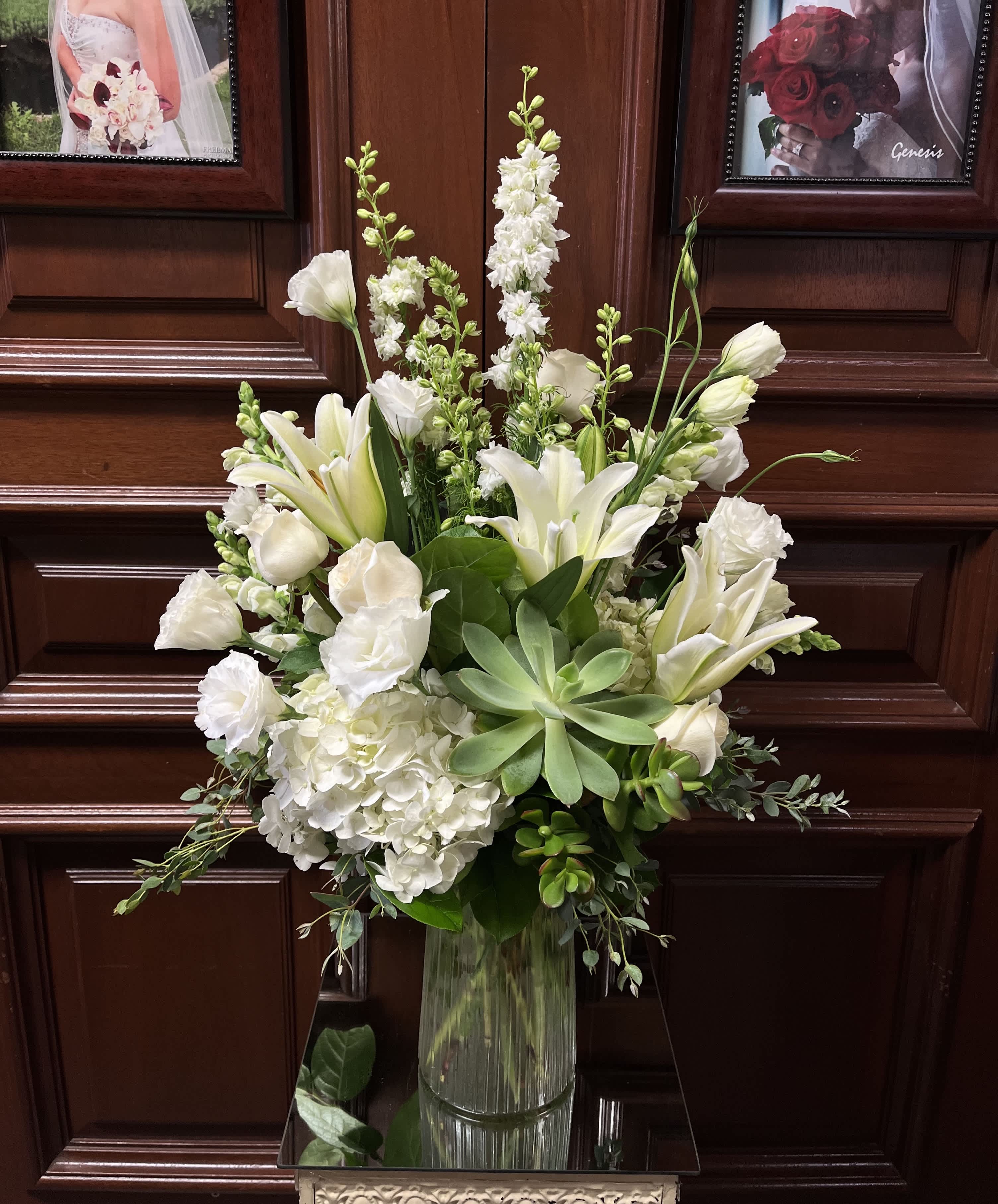 Opulent Serenity - Recipe: White Asiatic Lilies, White Delphinium, White Hydrangaes, White Roses, White Lisianthus, Succulent and greens in a tall, clear vase. Availability: All year round Design View: Symmetric Front Facing View Photo shown: Standard 