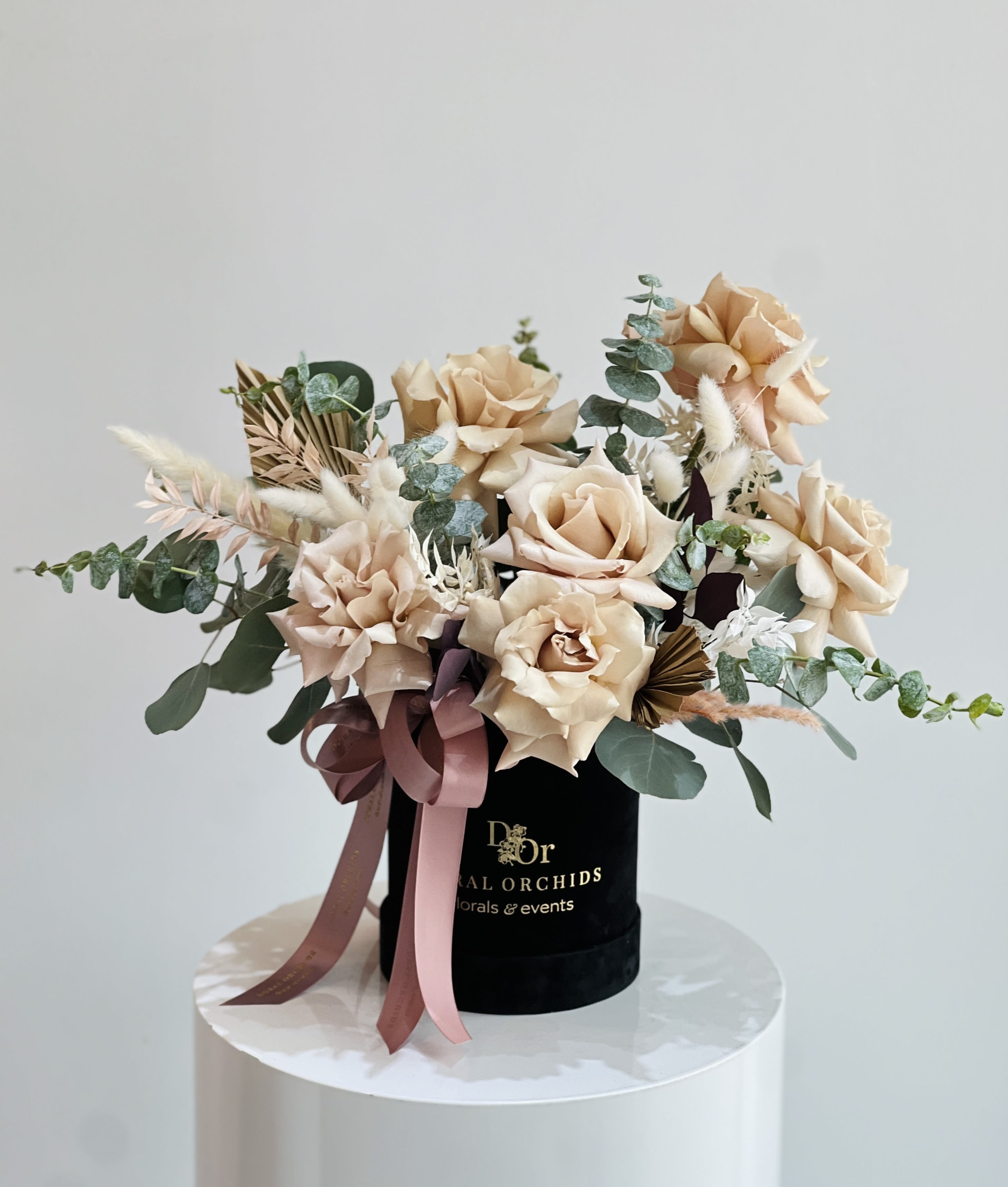 Gabriella™ - This arrangement features velvety, champagne-colored roses paired with airy eucalyptus and delicate dried accents for texture. Set in a sleek black velvet hatbox with flowing satin ribbons, it blends timeless romance with modern elegance.