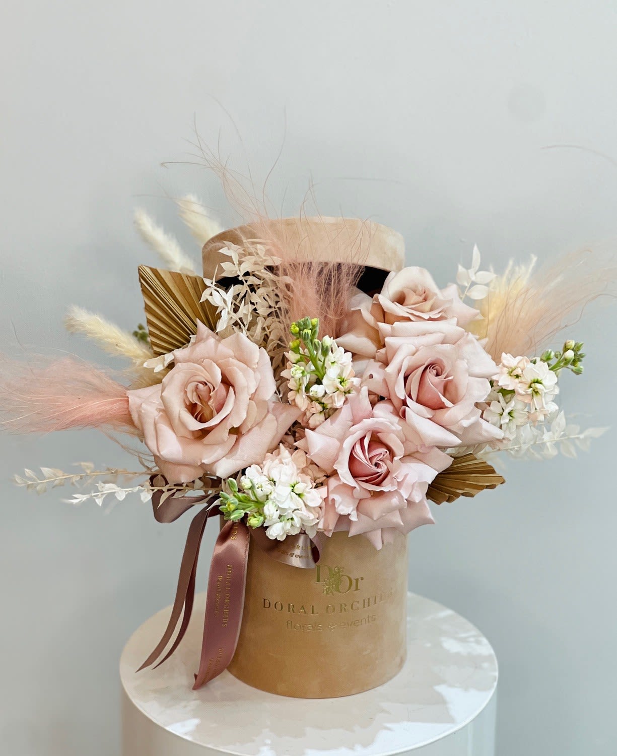 Sahara ™ - Velvety blush roses take center stage in this arrangement, softened by airy pampas grass, golden palms, and delicate white blooms. Presented in a suede hatbox with satin ribbons, it radiates romantic charm and refined elegance.