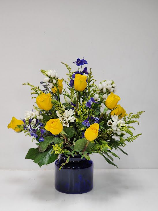 Magna Flora Magna Flora - Bright yellow roses, deep violet blooms, and delicate white flowers burst from a cobalt glass vase in this joyful, garden-inspired arrangement. Perfect for adding a pop of sunshine and elegance to any space. Approximately 20" x 18"