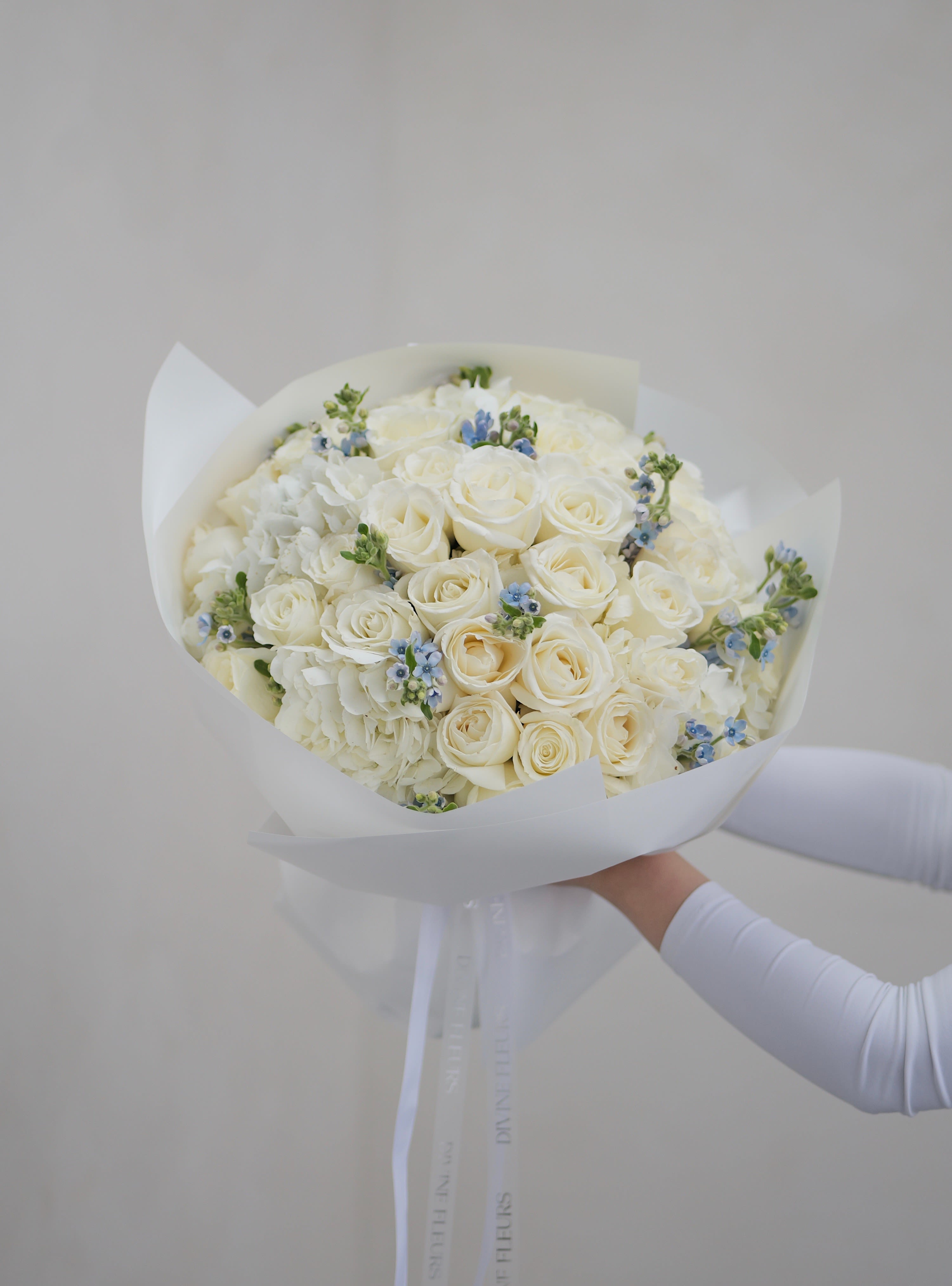 Touch of Blue - A soft and elegant bouquet of white roses and hydrangeas with delicate blue accents—perfect for welcoming a baby boy or celebrating a special moment with charm and grace. Wrapped in white and finished with our signature Divine Fleurs ribbon.  The bouquet pictured is shown in deluxe size.