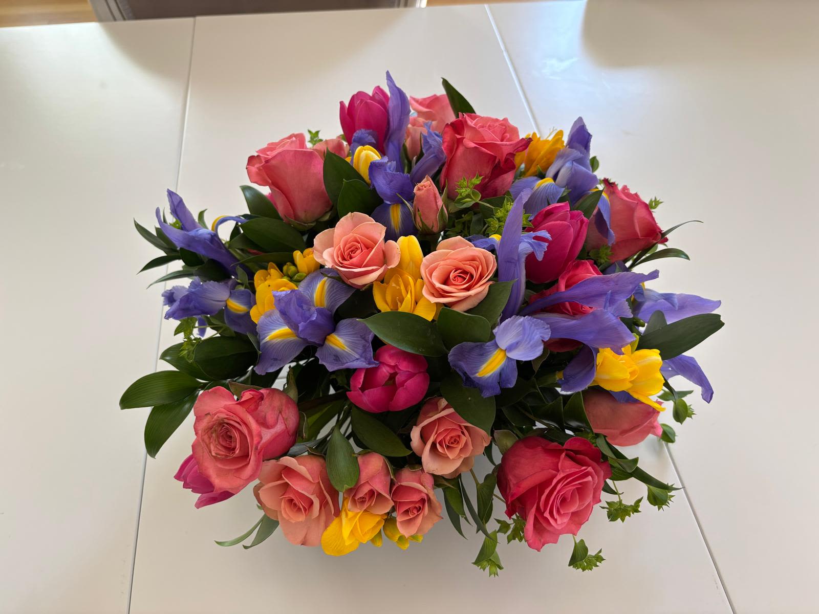 Joyful Symphony Joyful Symphony - A lively and vibrant floral arrangement that bursts with color and energy. Featuring delicate pink and peach roses, rich magenta tulips, sunny yellow freesia, and striking purple irises, all framed by lush greenery. This cheerful design brings a perfect harmony of elegance and freshness to any setting