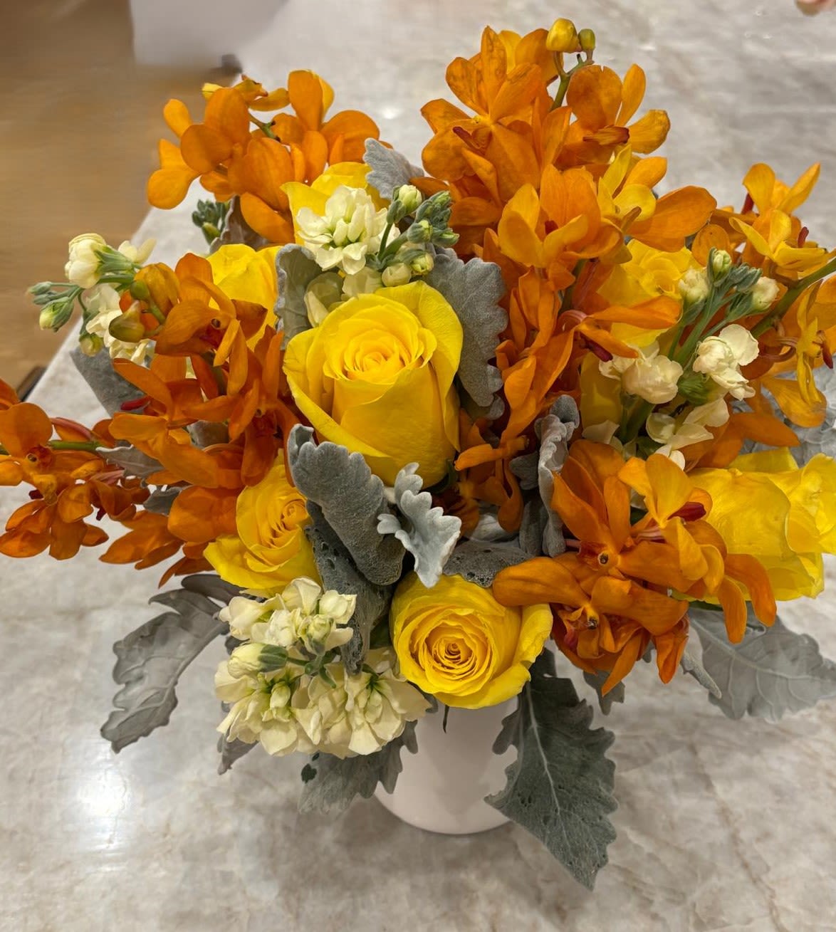 Golden Flame Golden Flame - A bold and radiant arrangement of vibrant yellow roses, fiery orange orchids, and creamy white stock softened with textured silver foliage. This striking design evokes warmth, energy, and elegance — the perfect statement piece to brighten any room or celebration