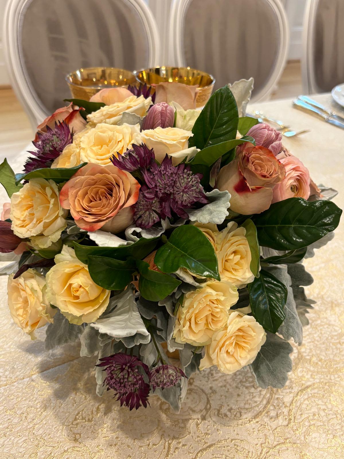 Golden Grace Golden Grace - An elegant and timeless floral arrangement blending soft peach and cream roses with rich burgundy accents and lush greenery. Complemented by silver-hued foliage, this refined composition exudes warmth, sophistication, and understated luxury — perfect for formal tablescapes or intimate celebrations