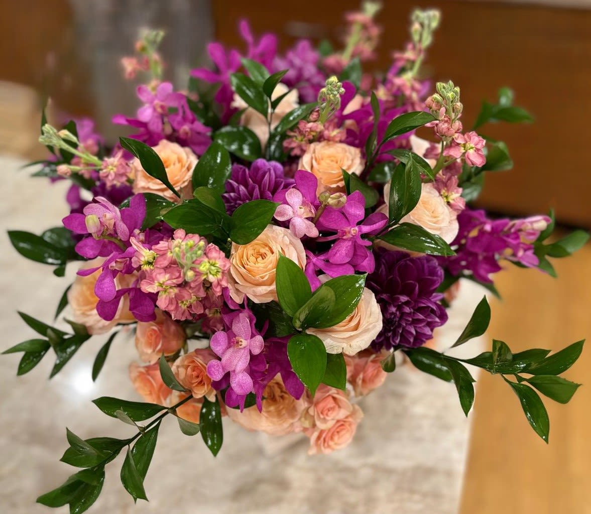 Orchid Sunset Orchid Sunset - An eye-catching, luxurious arrangement featuring vibrant magenta orchids, rich plum dahlias, and soft peach garden roses. Accented with delicate pink stock and glossy greenery, this bold design brings the warmth and drama of a tropical sunset to any elegant setting