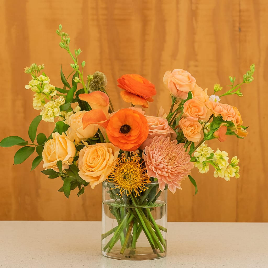 Golden Glow Golden Glow - A radiant mix of apricot roses, orange ranunculus, peach spray roses, and textured blooms like pincushion protea and mums, accented with fresh greenery. Designed in a clear glass vase, this cheerful arrangement brings warmth and sunshine to any room.