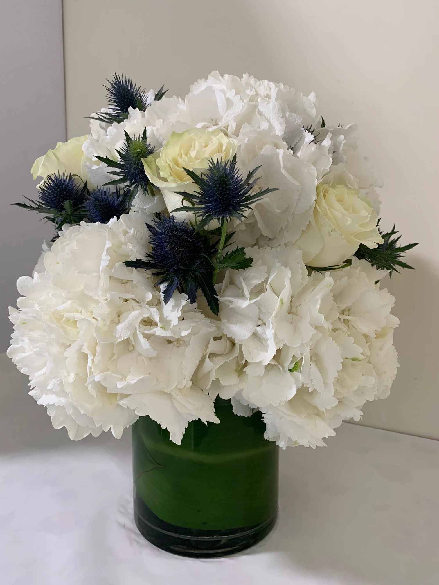 Southampton  - Dutch White - Introducing this amazing floral arrangement , made with the finest Dutch White Hydrangea and accented with White Roses and Euringuim  - Standing about 16 inches tall and 12-14 inches wide, this is designed in a 8 inch tall glass cylinder and wrapped with a Leaf