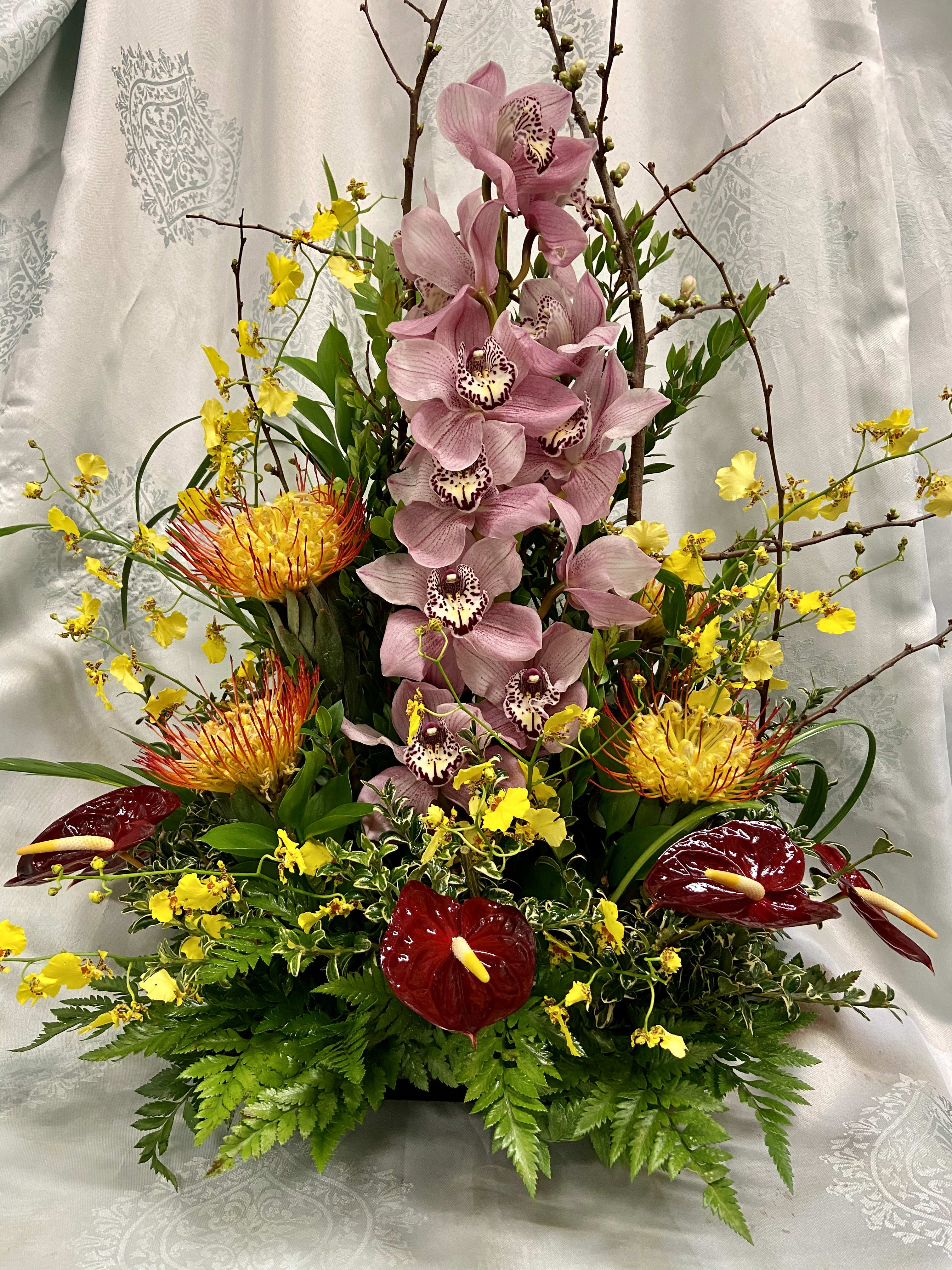 Tropical Hue - Tropical stylized arrangement using cymbidium orchids, proteas, anthuriums, oncydium orchid sprays with matching greenery and accented with Quince blossoms (subject to seasonal availability).