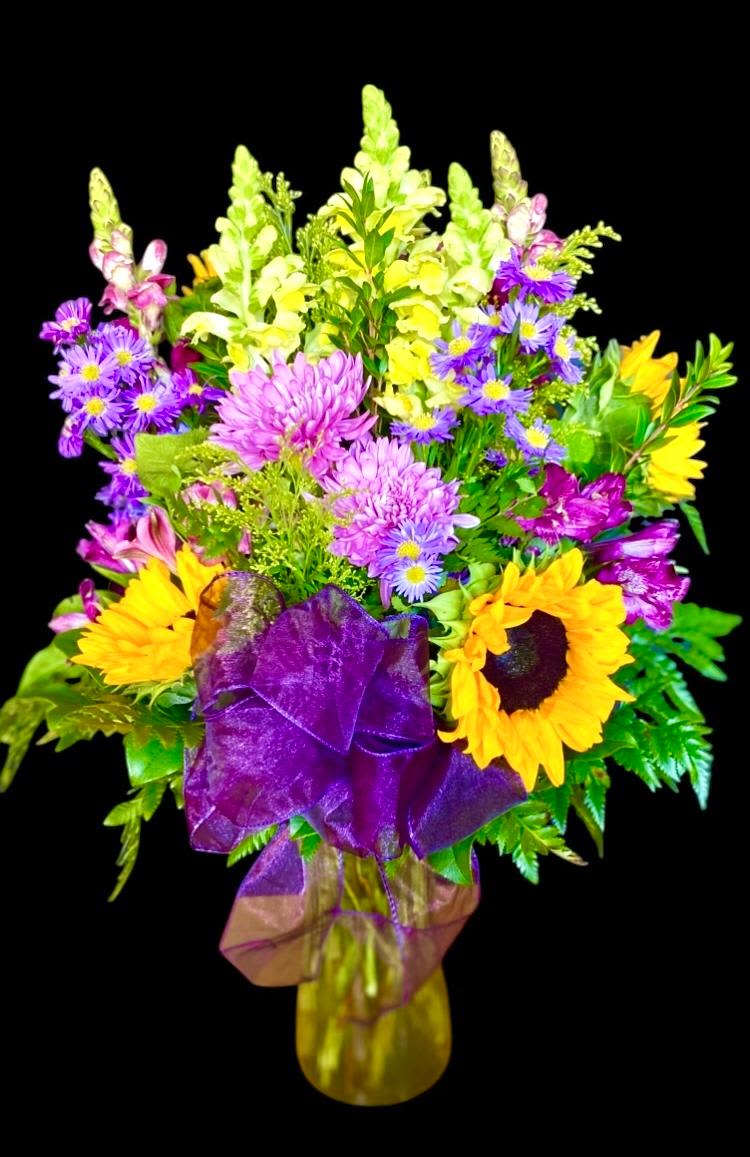 Cheerful Afternoon  - This bright warm design is composed of Sunflowers, China Mums, Alstroemeria, Monte Casino Asters, Snapdragon, and is accented with touches of Solidago. Set in various foliage in a clear glass vase and finished with a chiffon purple bow. This design measures approx. 12&quot;W x 18&quot;W.
