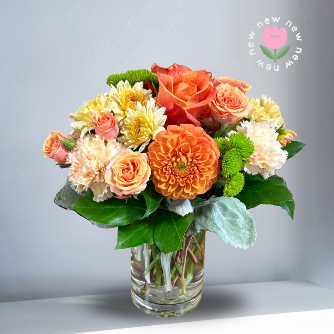 Summer zest  - Recipe: Seasonal dahlias, spray roses, orange roses, carnations, chrysanthemums, and dusty miller in a short clear vase. Seasonal: Seasonal dahlias Orientation: All Facing View Photo shown: Standard 