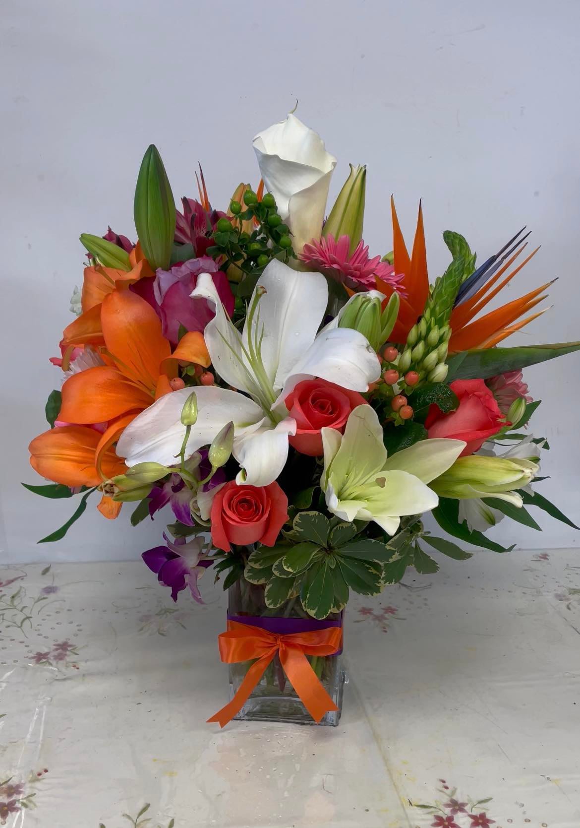 Tropical Land Tropical Land - A bouquet of mixed tropical flowers in bright colors in a square clear glass vase accented with a double satin ribbon. Cheerful and bright!