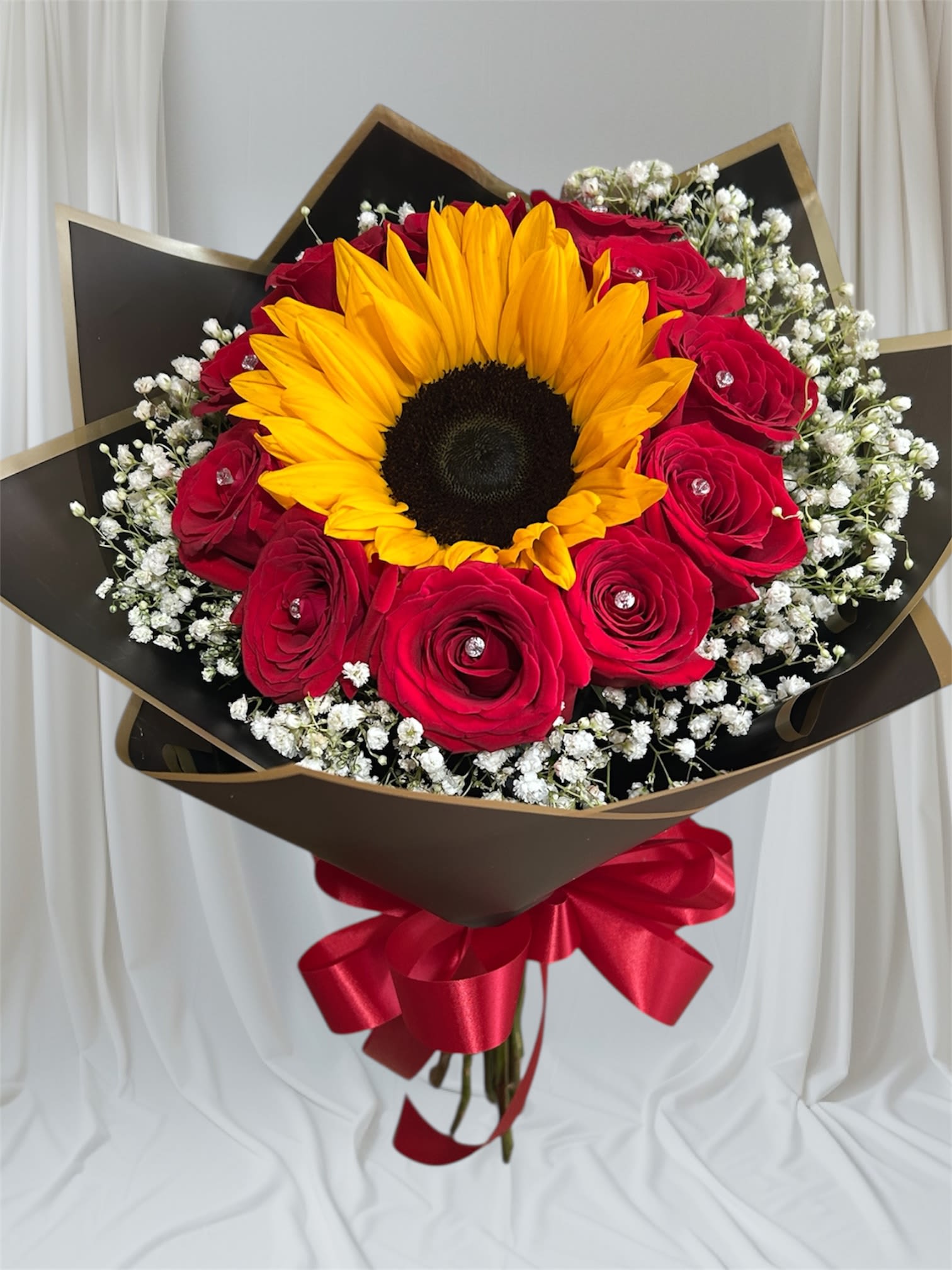 Sunshine Elegance Hand Bouquet - A radiant sunflower takes center stage, surrounded by lush red roses and delicate sprigs of baby’s breath. Wrapped in sleek black Korean paper, this bouquet blends romance and elegance with a touch of modern charm—perfect for making a bold and unforgettable statement.