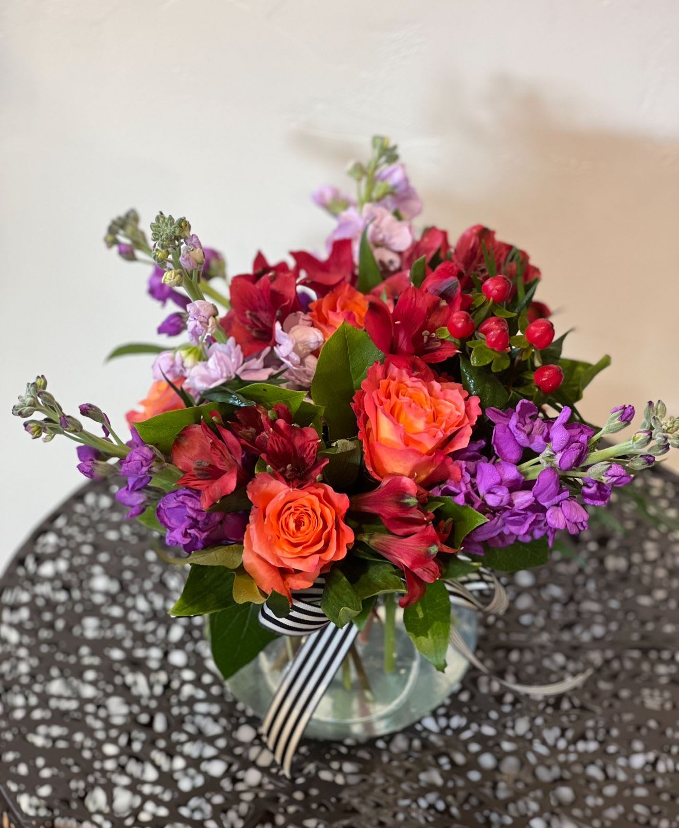 Rosy Posy - A fragrant jewel toned arrangement filled to the brim with stock, roses, alstromeria and hypericum in a glass vase.A perfect piece to set on an end table or coffee table. Measures approximately 9” tall. 