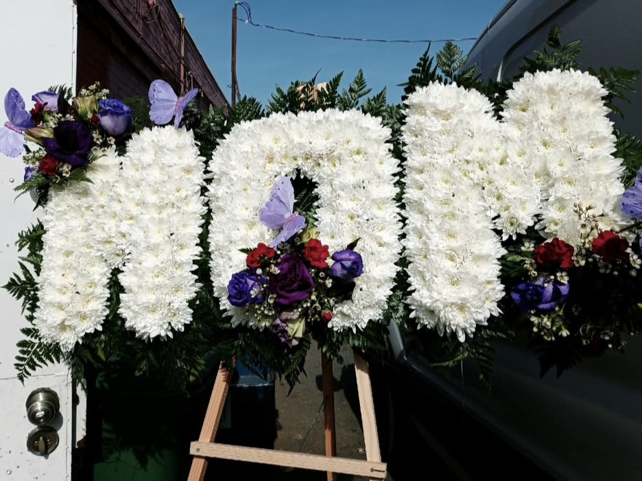 MOM #2 MOM #2 - This standing funeral tribute features the word ‘MOM’ in all white pompon cushions with a back layer of leather leaf. With corners and centers of blooming purple’s(roses, lisianthus, and wax) and a dash of red(carnations). The arrangement includes 4 decorative butterflies. Call us for custom change.