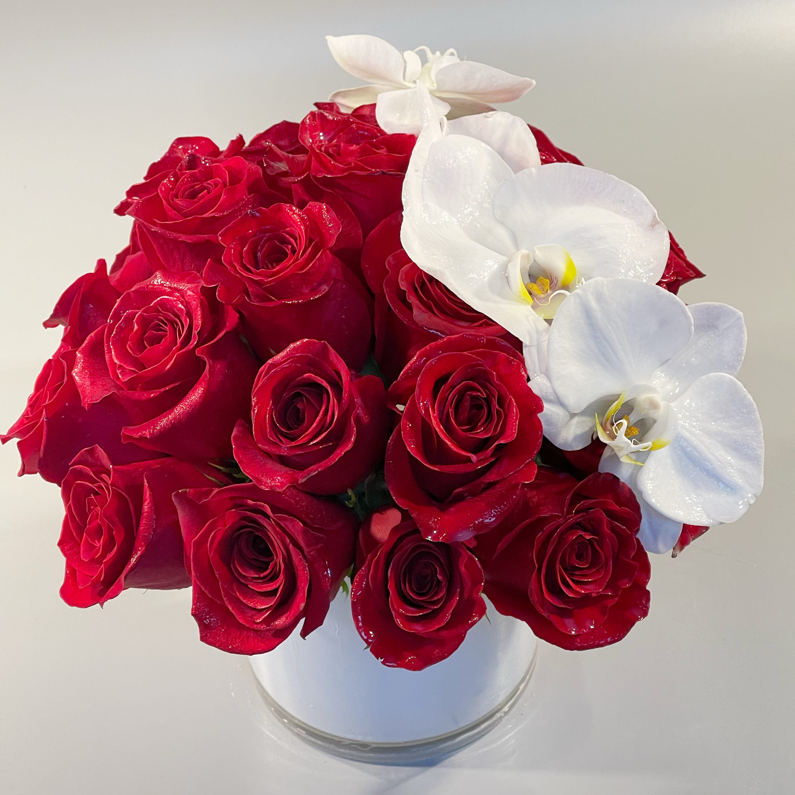 LOVING LOVING - A hand-tied French design arranged with red roses and orchid blossoms and placed in a white glass cylinder vessel.