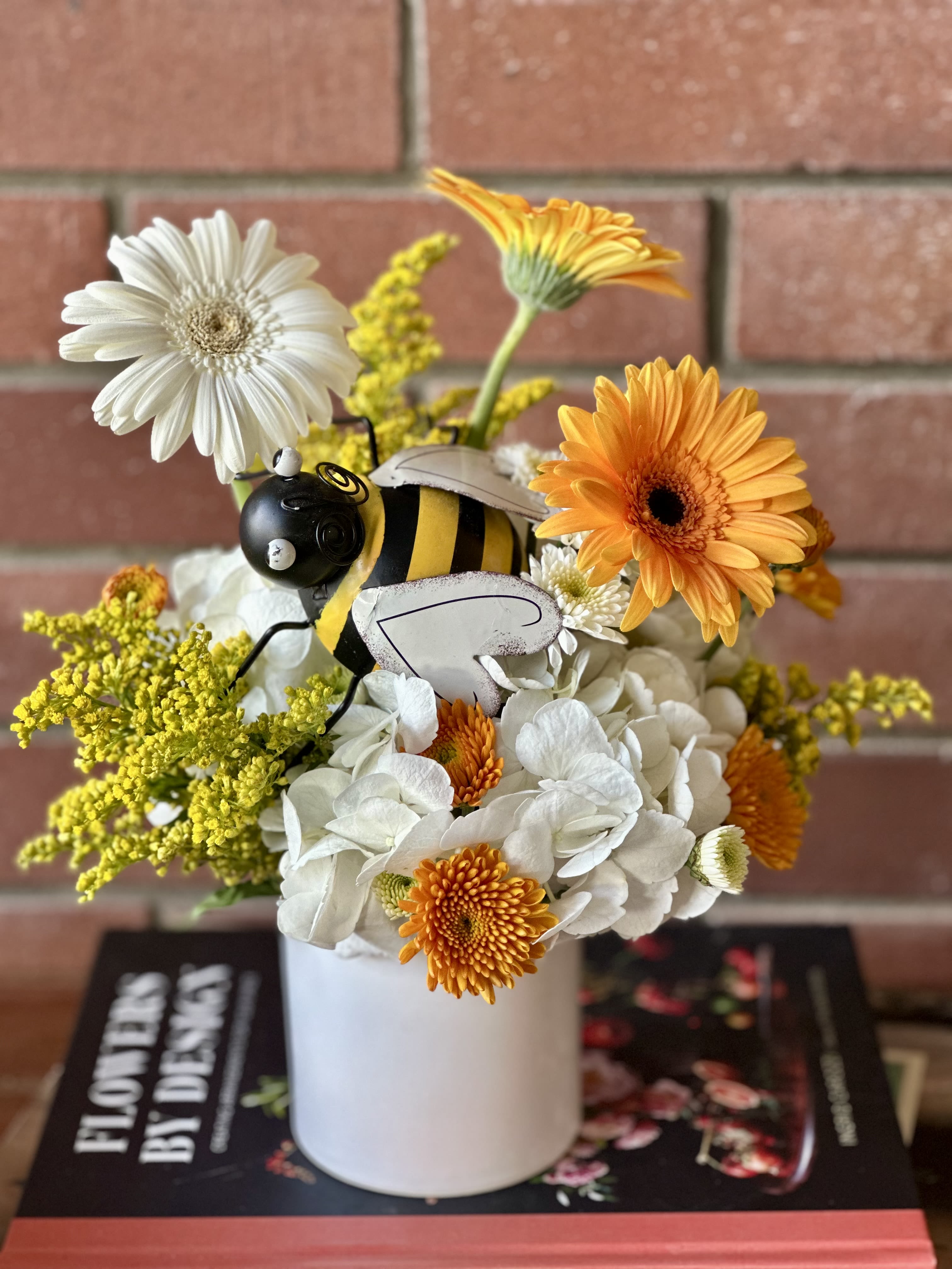 Bee Well Bouquet  - This cheerful arrangement captures the essence of a thriving garden with its vibrant mix of sunshine blooms and natural textures. Bright orange gerbera daisies take center stage alongside pristine white gerberas, creating a beautiful contrast that mirrors the energy of busy bees at work. Delicate white roses add romantic softness, while sunny yellow solidago (goldenrod) brings wild, meadow-like charm with its clusters of tiny golden flowers. The arrangement is thoughtfully accented with a whimsical bee decoration, complete with fuzzy texture and cheerful stripes, serving as a delightful reminder of nature’s hardworking pollinators. Presented in a clean white ceramic vase, this bouquet brings warmth and positivity to any space. Perfect for wishing someone well, celebrating recovery, or simply brightening someone’s day, the “Bee Well” bouquet combines the healing power of flowers with a playful nod to the industrious spirit of bees. Its sunny palette of oranges, yellows, and whites evokes feelings of optimism, energy, and natural wellness.