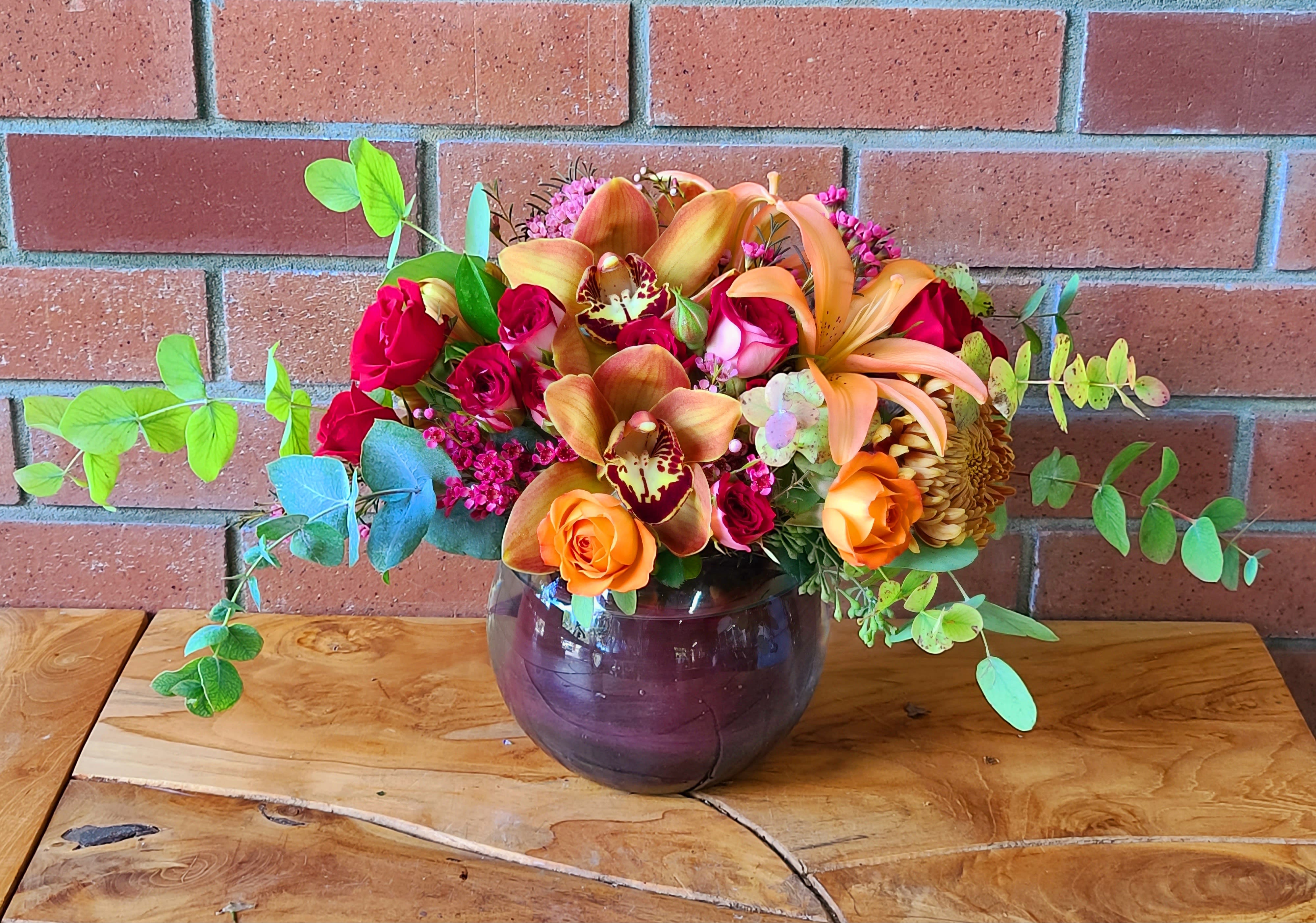Autumn Expressions Bouquet - Gorgeous fall theme arrangement in a clear bubble bowl wrapped with red ti leafs. Orchids, lilies, red spray roses, orange spray roses, lush eucalyptus and filler.