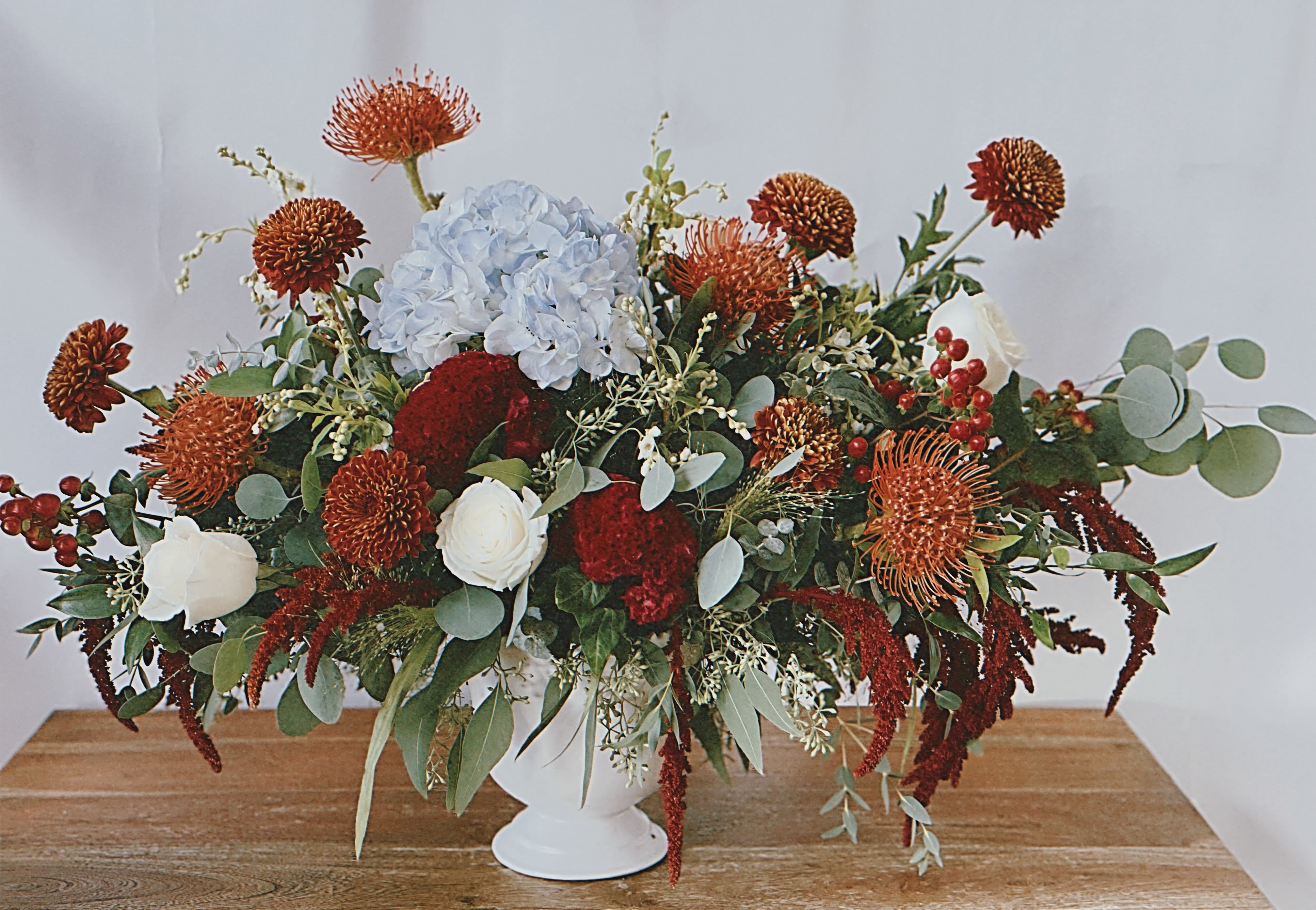 Classic Autumn Centerpiece  - The Classic Autumn centerpiece is a stunning arrangement that captures the essence of autumn with a vibrant and fresh aesthetic. Dominating the composition are gorgeous orange pin cushion proteas, their unique shape and vivid hue adding a striking focal point. Surrounding them, clusters of cheerful orange mums bring warmth and a sense of celebration, harmonizing perfectly with the proteas.  Interspersed throughout the arrangement are tropical celosia flowers, their feathery texture and rich color providing an exotic flair. The elegant white garden roses add a touch of sophistication, their soft petals contrasting beautifully with the bold oranges. Complementing this colorful display is the deep blue hydrangea, which introduces a cool tone that balances the warmth of the oranges and creates visual interest.  The centerpiece is further enhanced by upscale fall greenery and filler flowers, which provide a lush backdrop and add depth to the arrangement. The combination of textures and colors creates a dynamic and eye-catching centerpiece that embodies the spirit of a fall fiesta, making it perfect for any festive gathering. Crafted by Venetian Flowers, this arrangement not only showcases the beauty of the season but also elevates the overall ambiance of the event.
