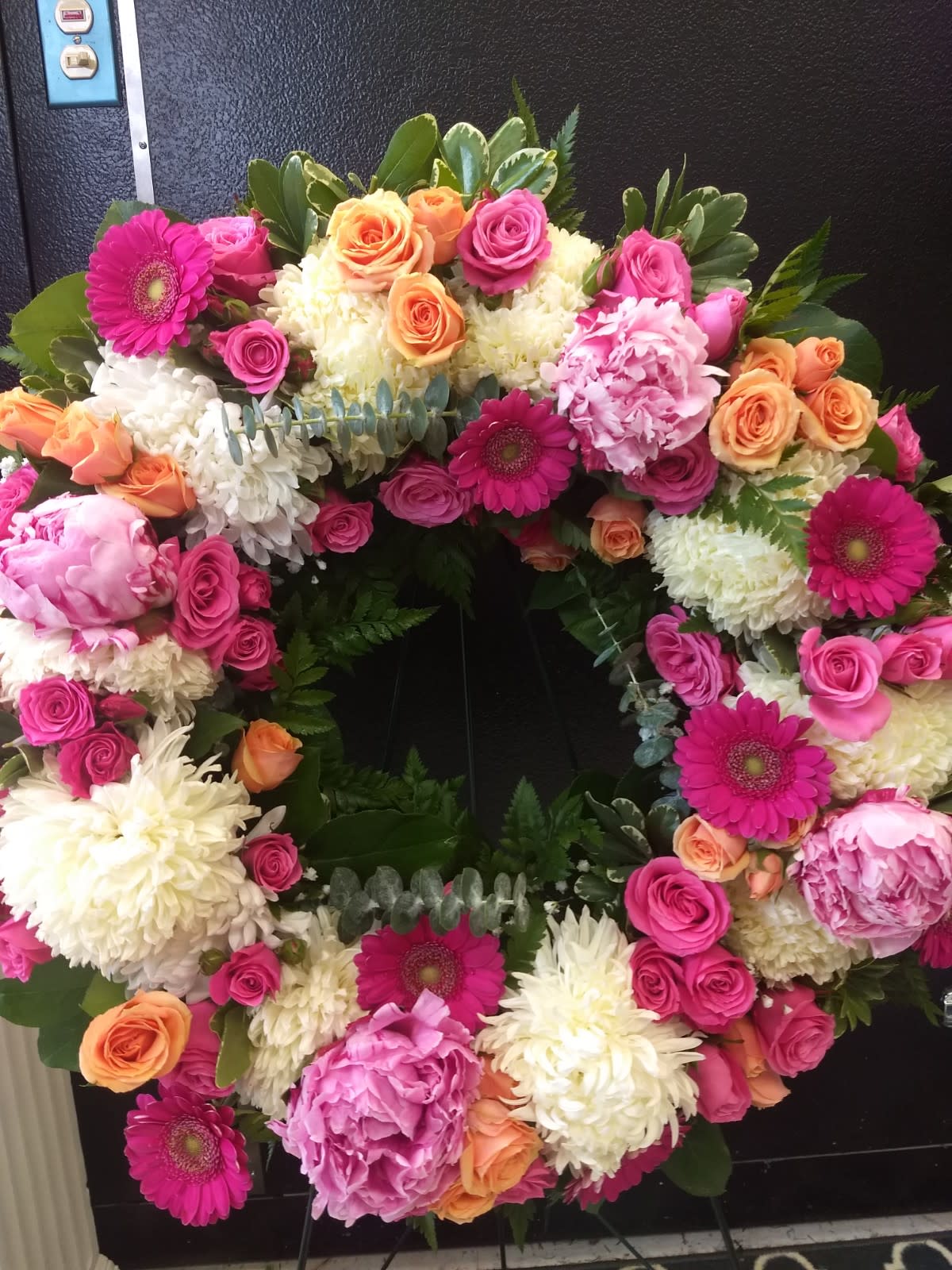 Grace, bright wreath - Honor with a beautiful wreath composed of our bright mix florals including lilies, peony, gerbera,  mums and roses.  **** Please note: 48 hour notice MAYBE REQUIRED We custom-design this arrangement using best-of-day seasonal flowers. This image is a general representation of its size and style and may not feature the exact flowers shown. Due to disruptions in the global supply chain and staff shortages, we may have to make last minute substitutions to your selected design. If we need to make significant changes we will call you first to get your approval*** Thank you for understanding.  ******Due to disruptions in the global supply chain and shortages, we may have to make last minute substitutions to your selected design. Don't worry, it will be beautiful! If possible on Sympathy orders to the funeral home 24 to 36 hour notice is best, so that we can try and make sure we can source your flowers. Thank you for understanding. if we need to substitute product that will alter the feel of the design, we will contact you before sending it out.****