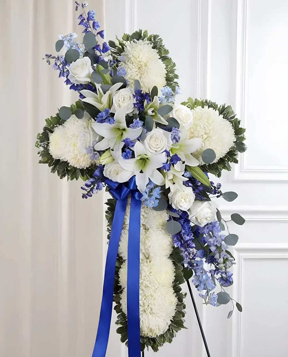 Cross White &amp; Blue - This cross-shaped arrangement offers a graceful symbol of faith and remembrance. Designed with fresh blooms, it brings comfort and hope during times of loss. Placed on an easel, it stands as a heartfelt tribute.