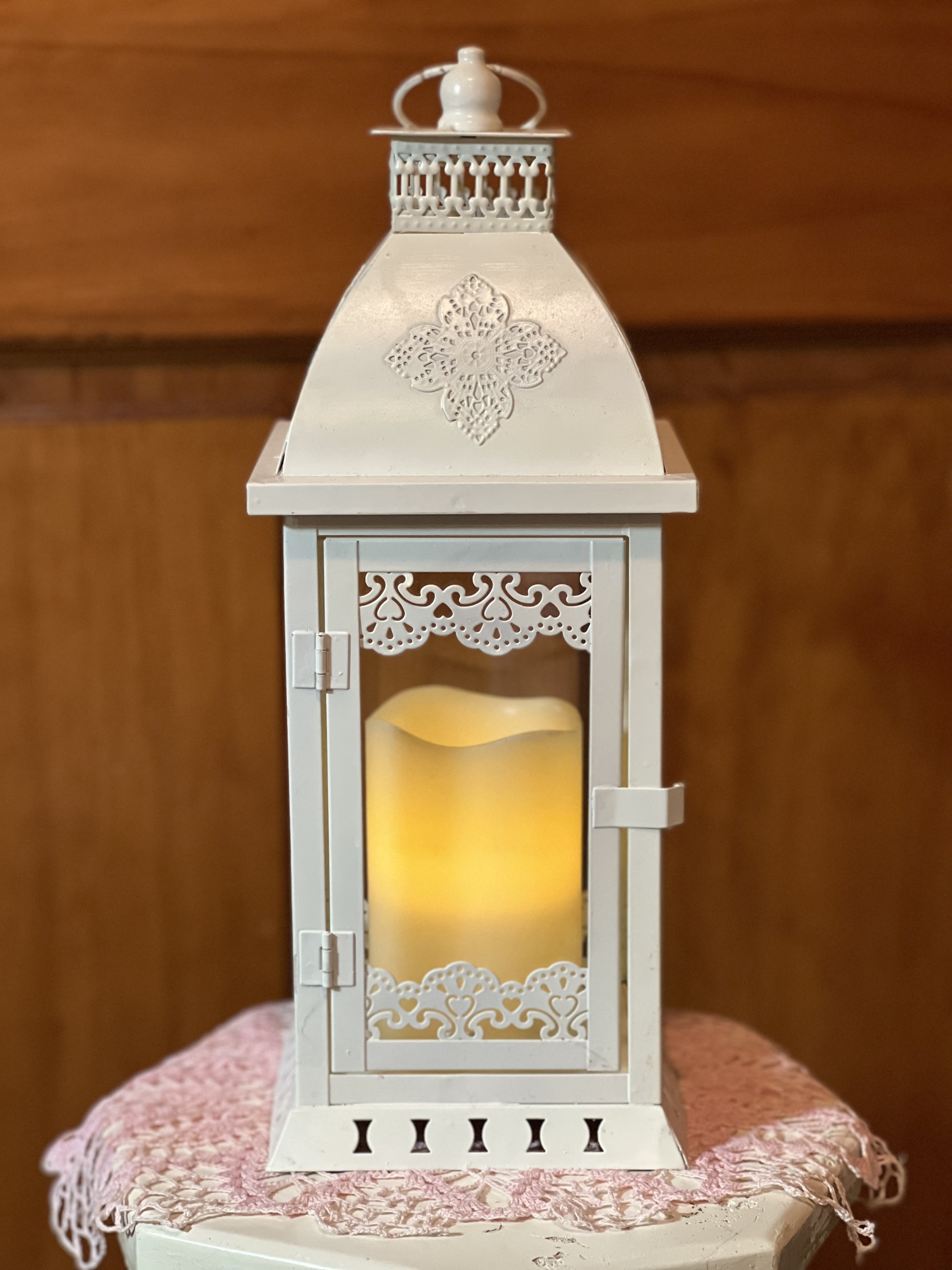 Pillar Lantern Pillar Lantern - A Beautifully metal textured off-white 14" tall lantern fitted with glass panes included with a flameless LED candle (3" X 5"). 2 AA batteries included. Candle made of paraffin wax. FOR INDOOR USE ONLY. Do not place near any heat source that could cause wax to soften or melt. Safe, no-flame, no fire hazard, no drips, no mess. Adding a warm light and style to any room in your home. Candle shown lit. ( DELUXE: We will add in a base, surround with seasonal flowers, and greenery in a waterproof container.)