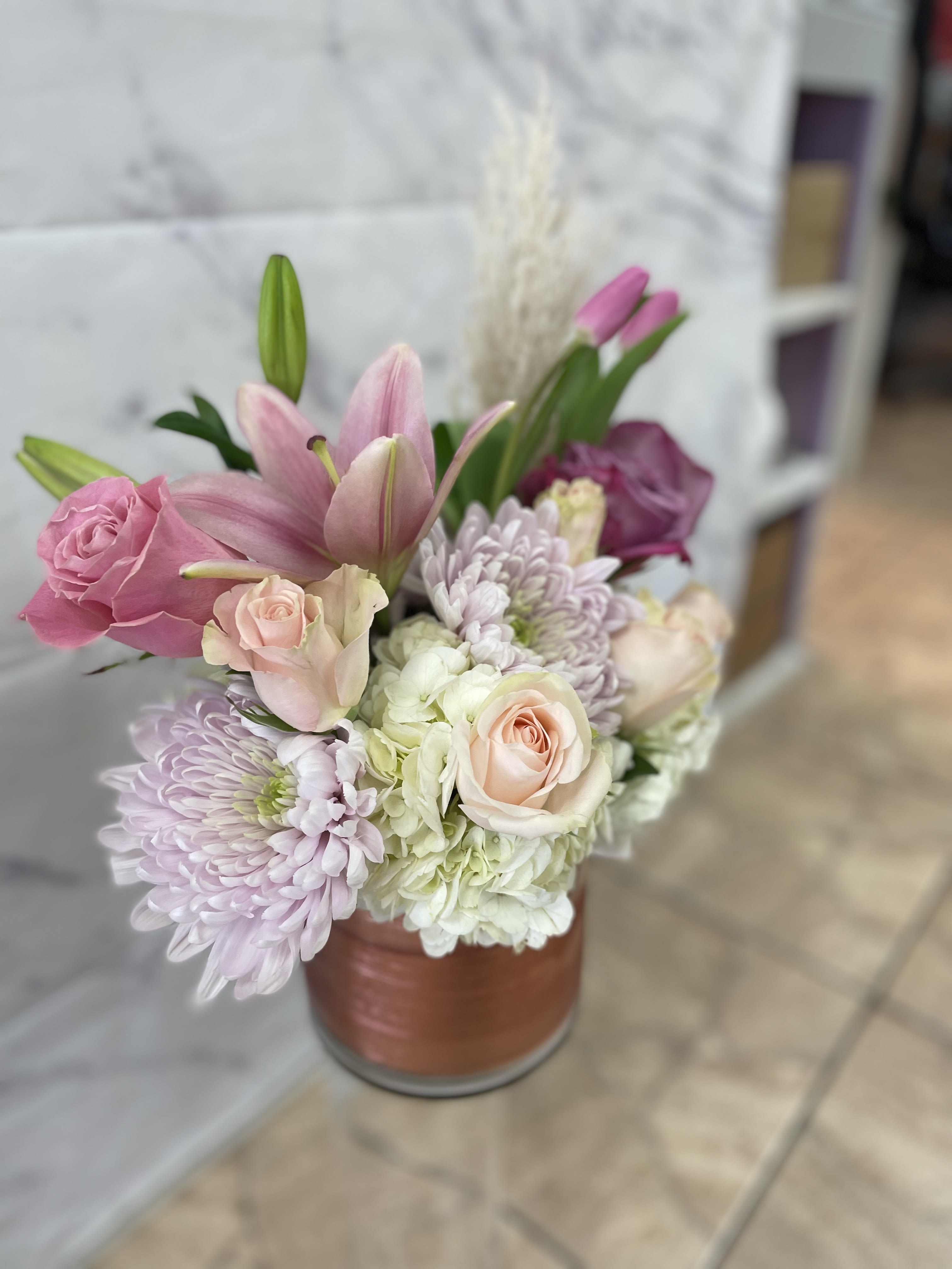 Modern Mom - A chic cylinder with a pink wrap in the vase blooming with a mix Of lush flowers And a hint of pampas grass