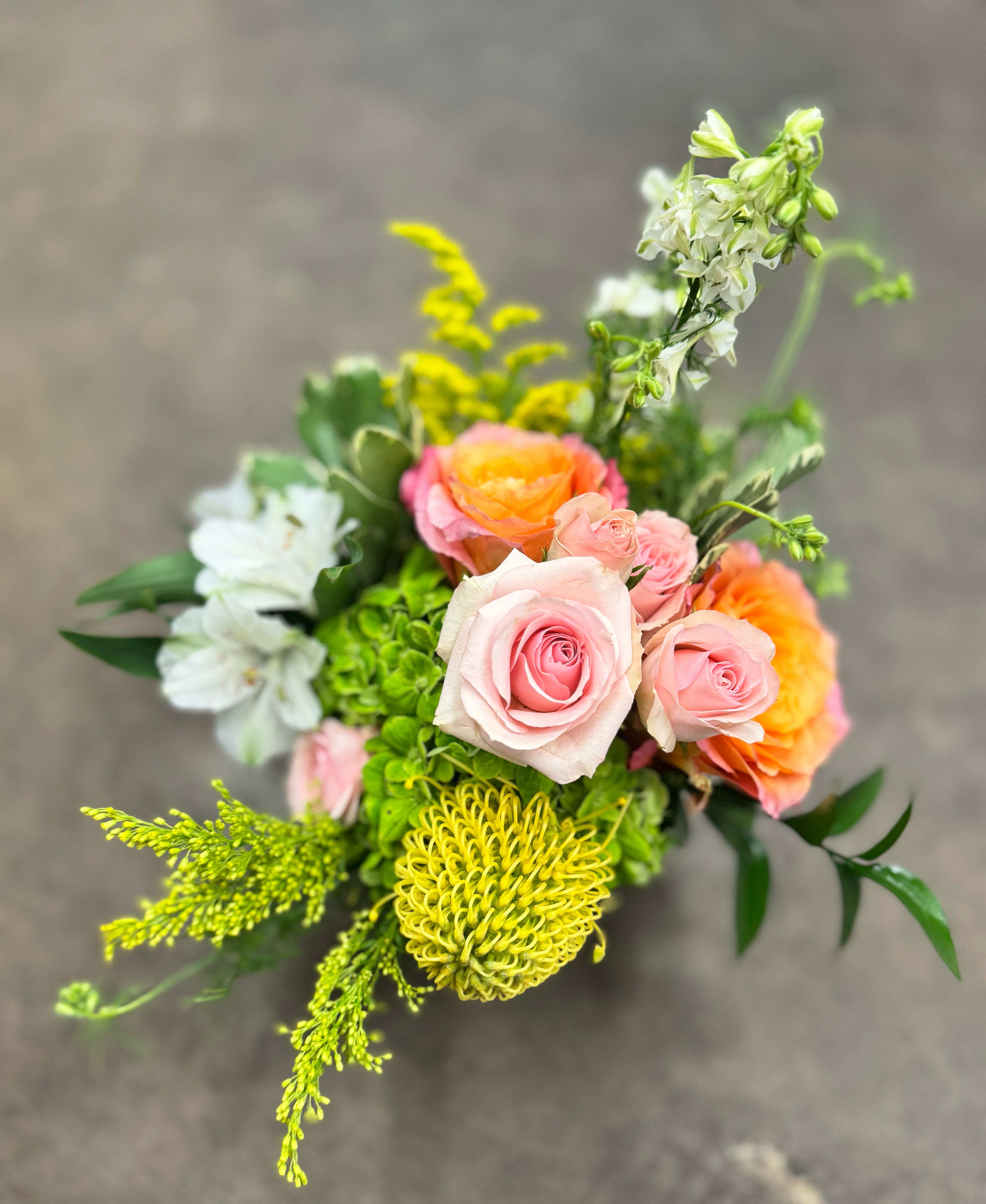 Citrus Garden - Bright and cheerful, this garden-style design pairs orangey/peach roses, pink spray roses with lime-green hydrangea, white alstroemeria and larkspur, golden solidago, and a sunny pincushion protea. Styled with fresh greens in a clear glass cube, it’s a vibrant pick for birthdays, thank-yous, or just because. Hand-delivered by Thompson’s Flower Shop with same-day service in Goodyear, Litchfield Park, Verrado, PebbleCreek, and nearby.