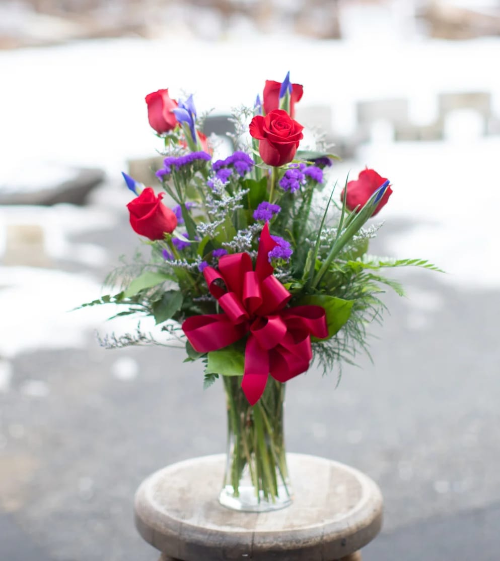 Stunning Beauty - A beautiful all around vase showcasing six elegant crimson roses with six spring indigo iris flowers. Seasonal foliage and purple statice complete the design displaying a similar color tone as the iris flowers before they open. Your recipient will enjoy watching the iris slowly open as they acclimate. The iris flower has a kiss of sunshine yellow, contrasting perfectly with the indigo blue hues or the bulb bloom. 