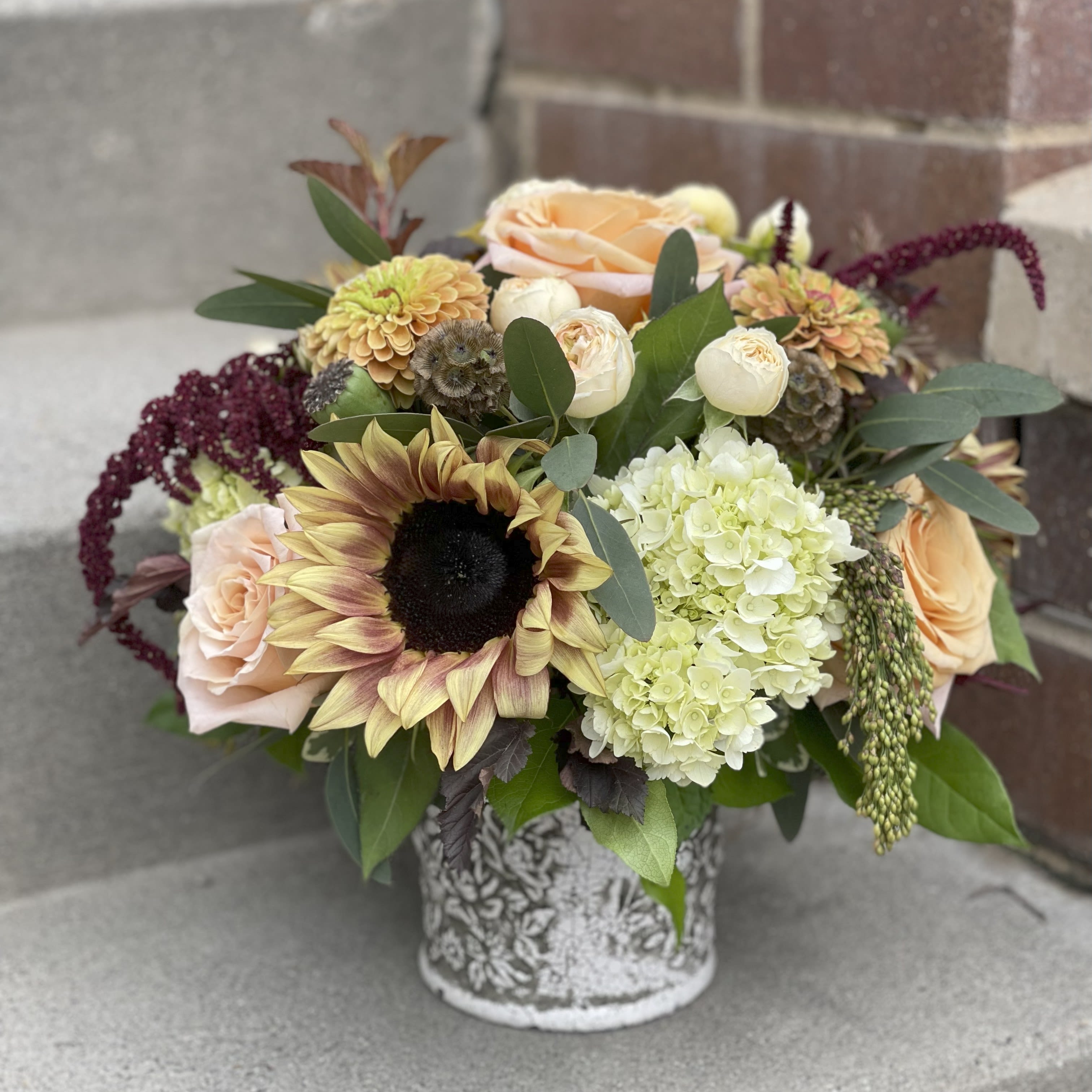 Autumn Arrival - The perfect addition to any autumnal décor. This stylish and compact arrangement features a mix of the local blonde sunflowers, mini green hydrangea, Amaranthus, and seasonal local zinnias, all arranged in a rustic stoneware container. The warm and vibrant hues of this centerpiece perfectly capture the essence of the season, making it a delightful display for any home or event. With its elegant design and locally sourced blooms, the Autumn Arrival is the ideal way to bring a touch of nature indoors during the fall months or herald the end of summer. 
