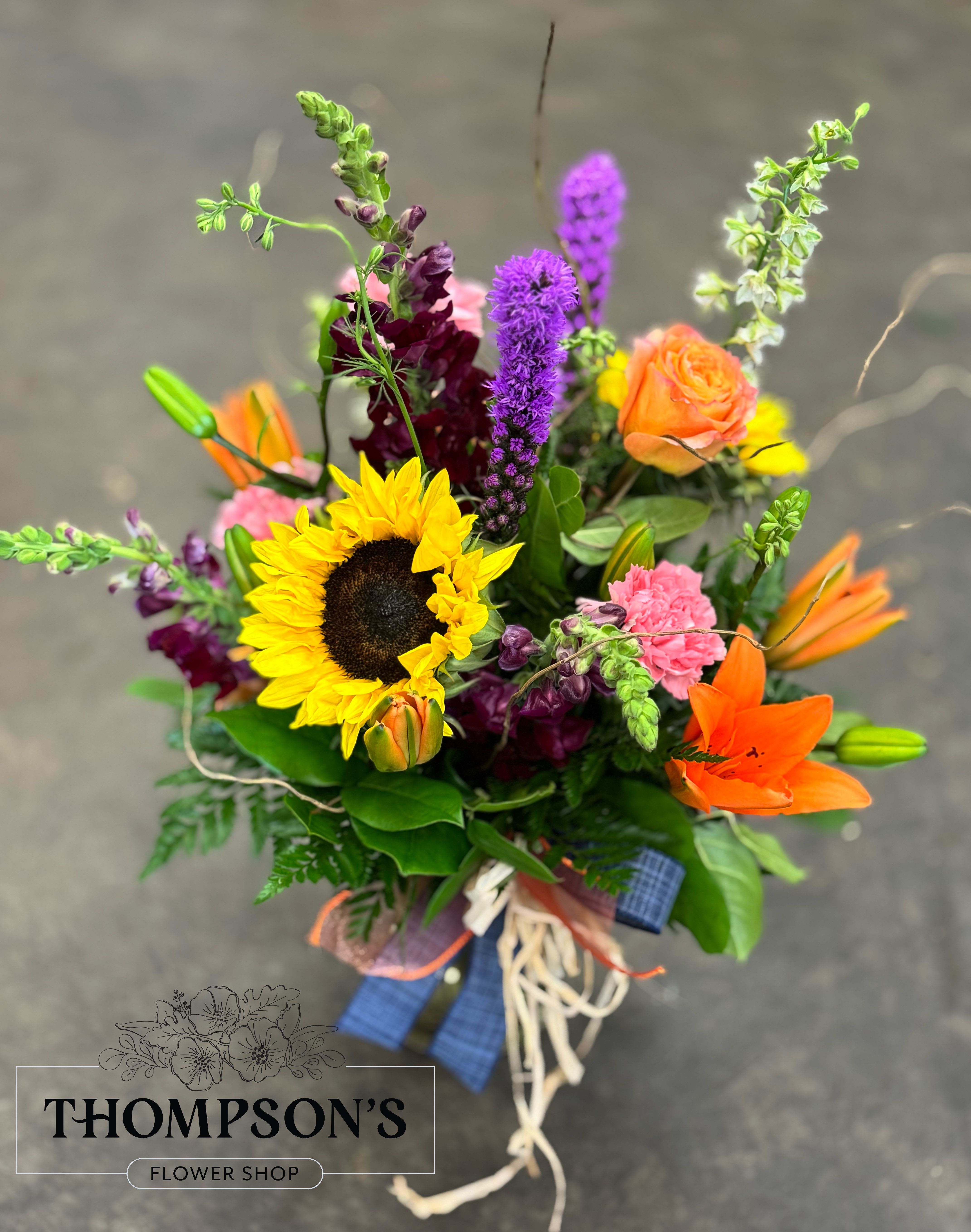 Sunrise - Looking for a taste of fall colors in the end of the summer season? This mix of sunflowers lilies, snapdragons and curly willow are the perfect mix of summer and fall colors!  This is a round arrangement with flowers all the way around. 