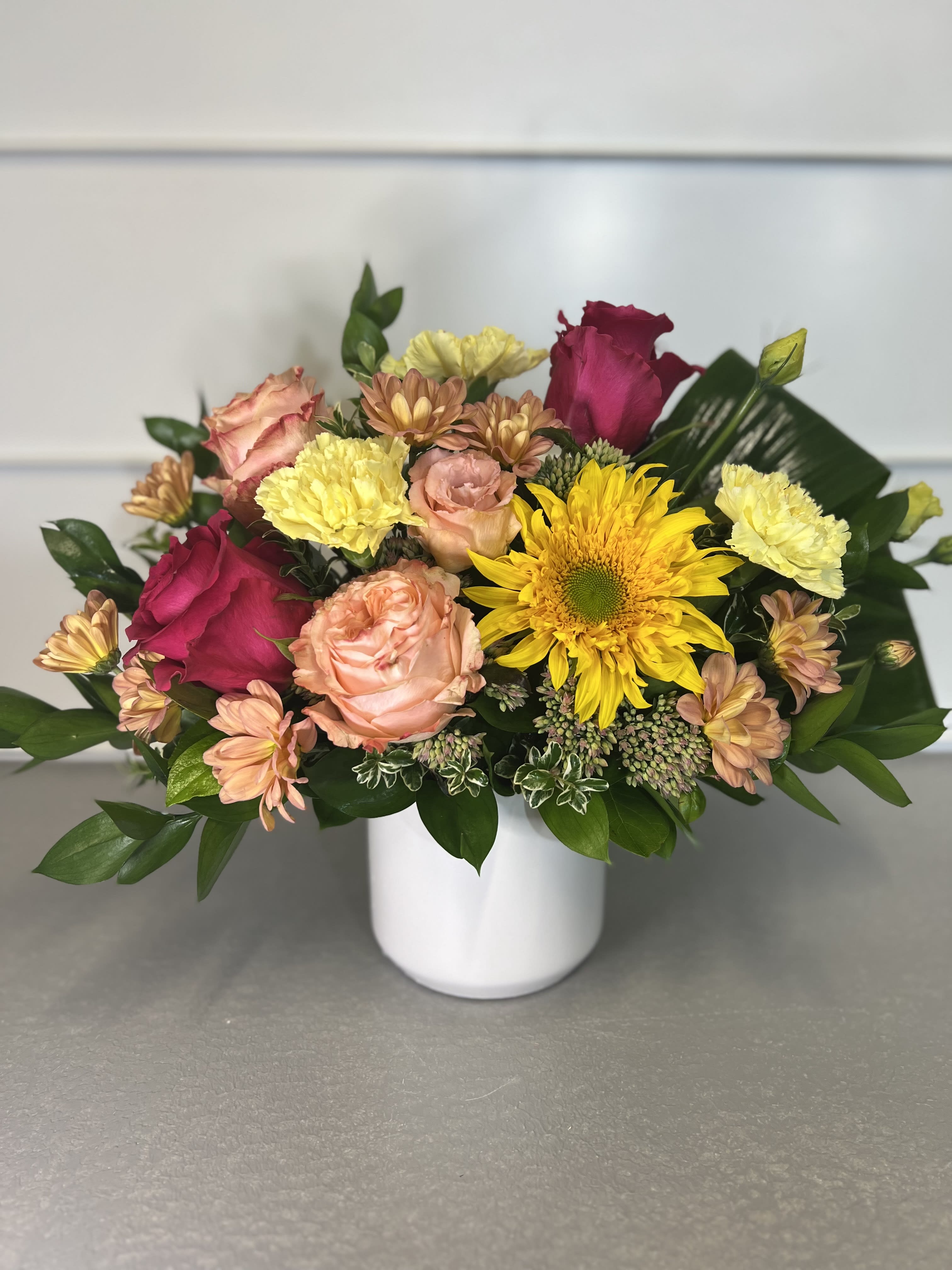 Alicia  - This bouquet includes mixed greenery, roses, gerbera daisies, carnations, chrysanthemums, and lisianthus in a white ceramic pot. 