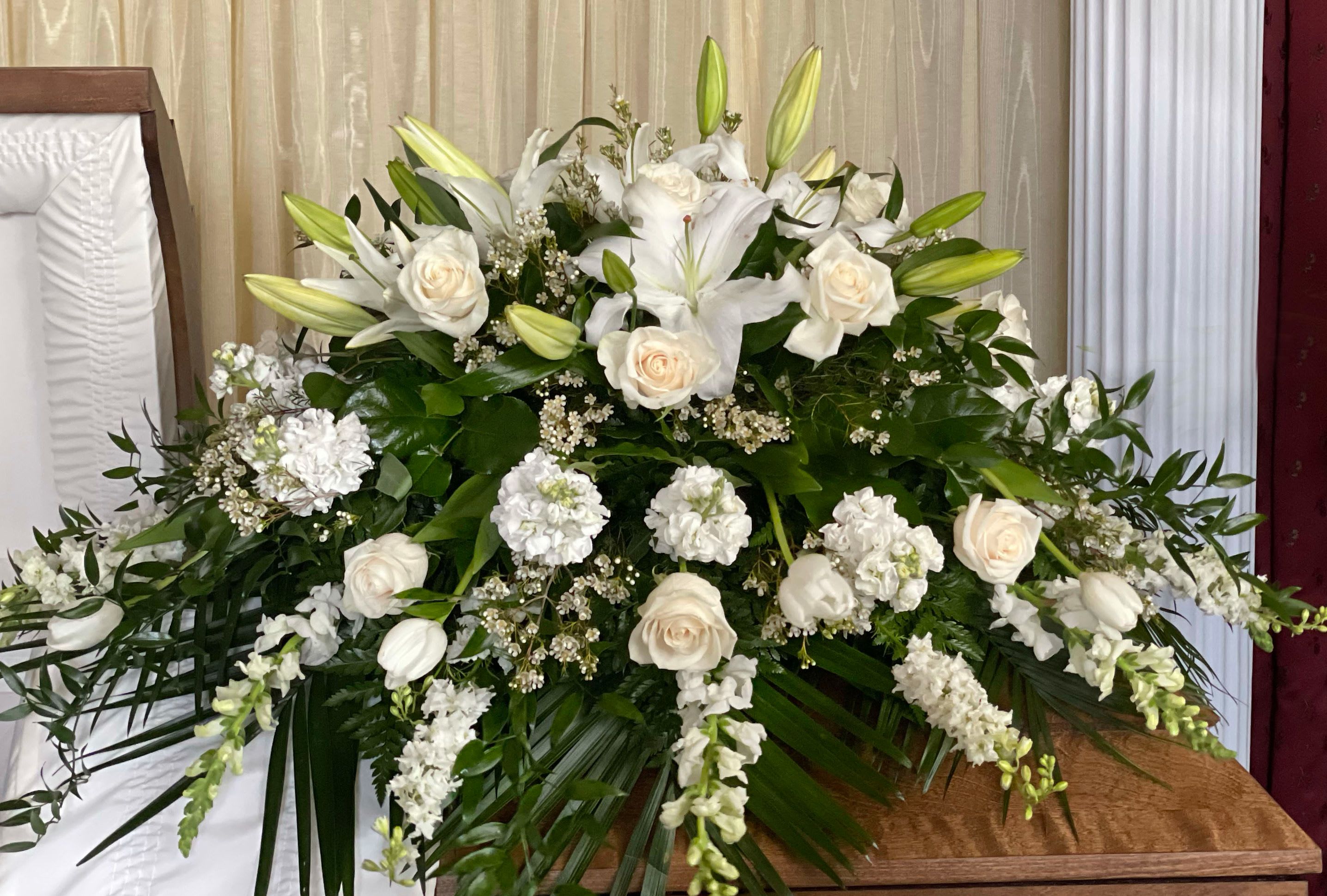 Casket Spray CS-32 - This refined casket spray offers a graceful farewell with its soft blend of white lilies, roses, snapdragons, and seasonal white blooms. Accented with fresh greenery and flowing textures, it conveys peace, reverence, and unconditional love. A timeless expression of remembrance, perfect for honoring a life marked by dignity and kindness.