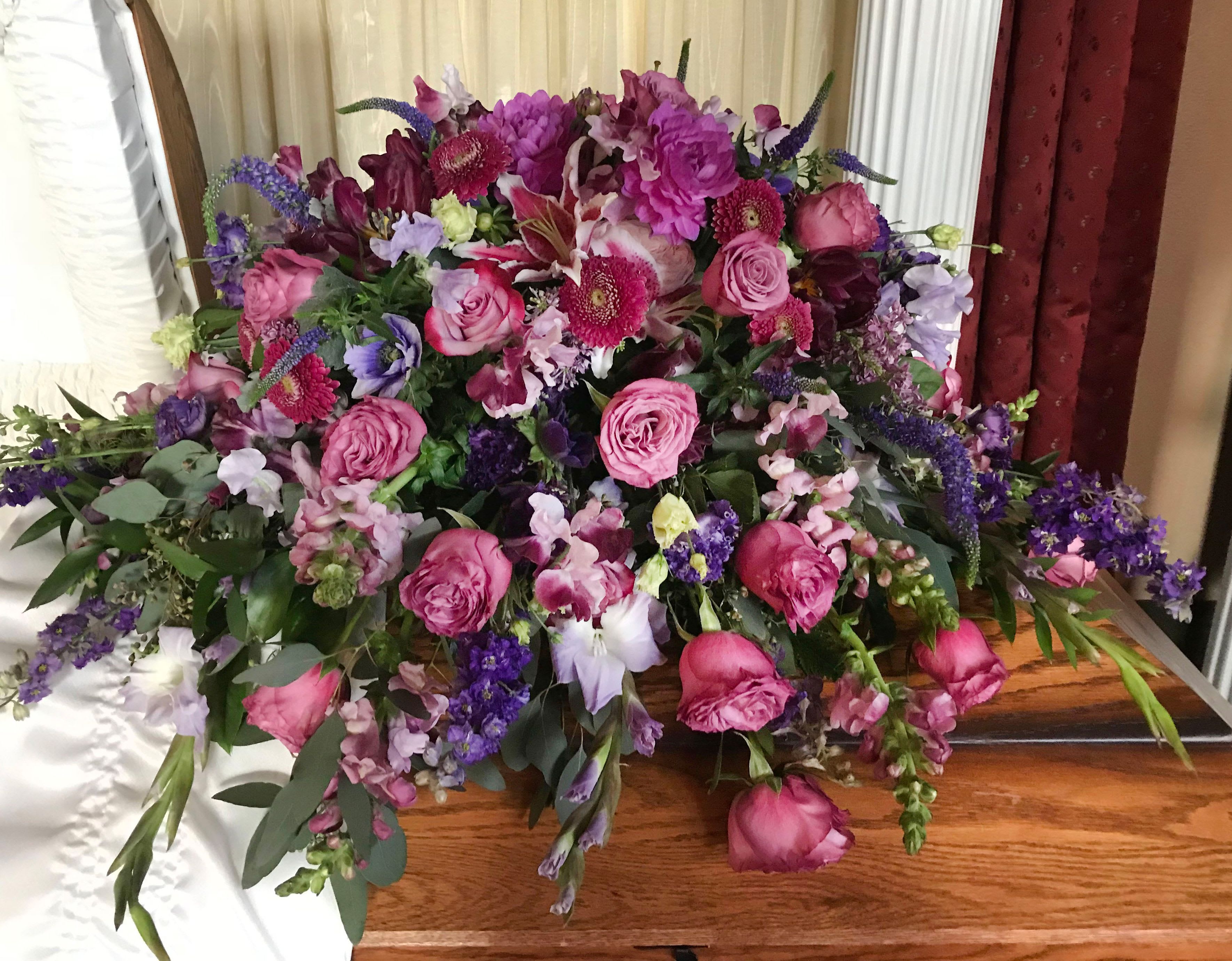 Casket Spray CS-12 Casket Spray CS-12 - Overflowing with soft elegance and delicate beauty, this casket spray features a lush blend of lavender and pink roses, snapdragons, purple larkspur , stock, orchids, veronica, and seasonal garden blooms. Hints of eucalyptus and cascading textures add natural movement and depth, creating a graceful and heartfelt tribute. Designed to rest gently atop the casket, this arrangement honors your loved one with a peaceful palette and a garden-inspired feel—perfect for celebrating a life lived with kindness, beauty, and love. Customization is available—please contact us if you’d like to personalize the color scheme or flower selection to reflect your loved one’s memory.