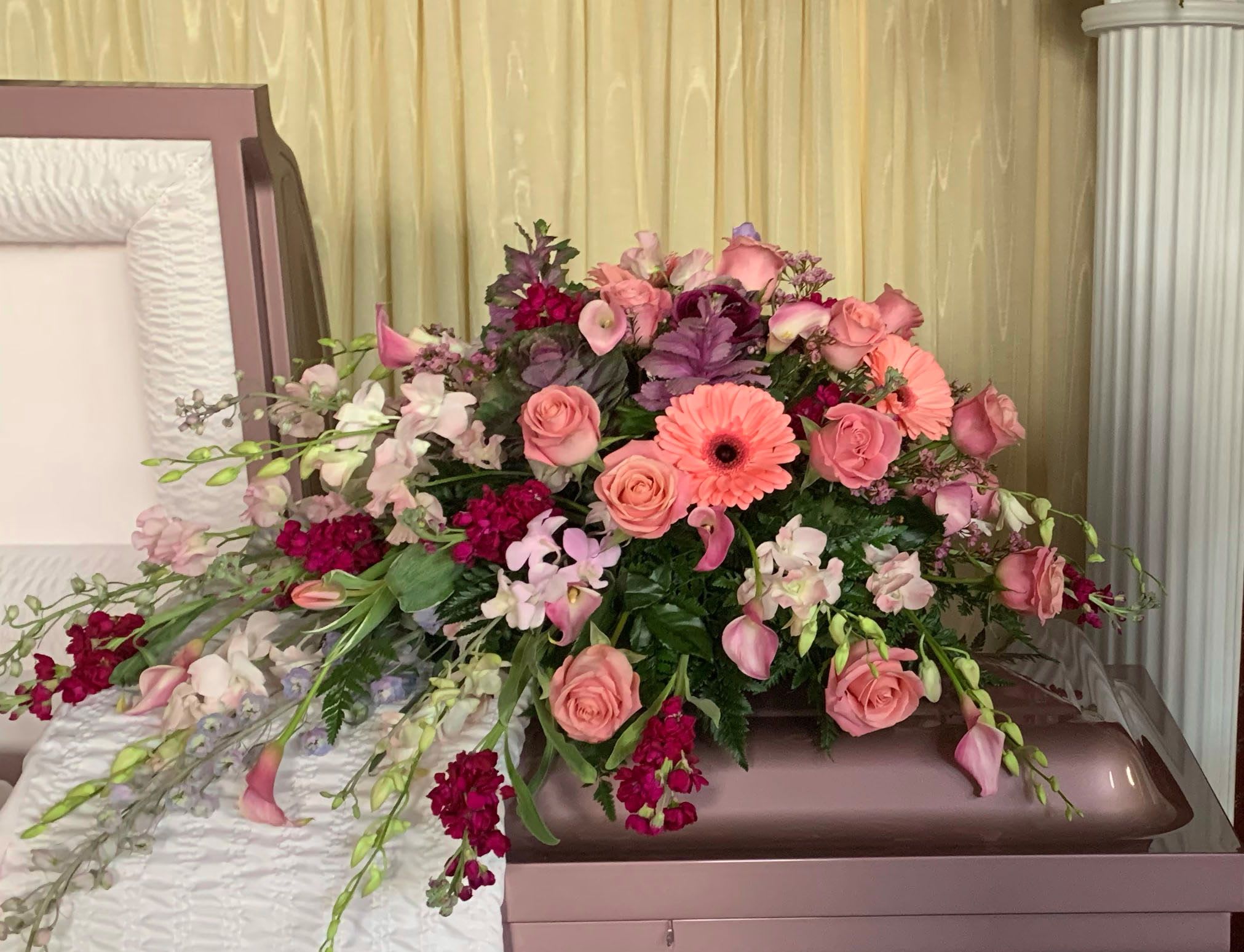 Casket Spray CS-13 - A soft and heartfelt farewell, this casket spray features a soothing blend of peach and pink roses, gerbera daisies, mini calla lilies, snapdragons, orchids, and rich magenta accents. Designed in graceful layers with flowing greenery, this arrangement conveys warmth, gentleness, and deep love.  Perfect for honoring a mother, daughter, or cherished friend, this feminine casket spray brings beauty and comfort to the service with its romantic color palette and garden-inspired design.  If you wish to customize the flower selection or color scheme, please contact us—we're here to help create a tribute that feels personal and meaningful.