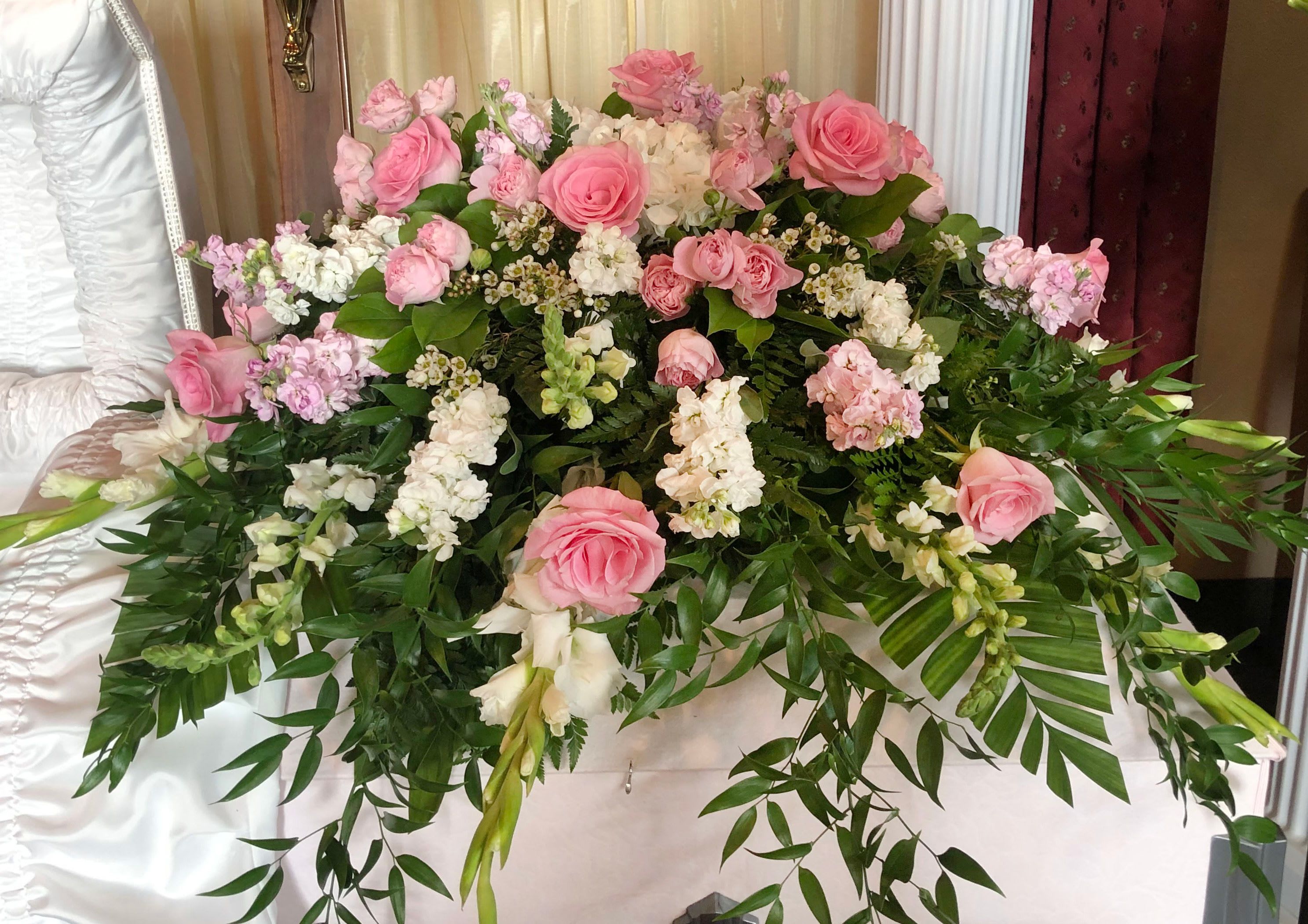 Casket Spray CS-14 Casket Spray CS-14 - Soft, tender, and peaceful, this elegant casket spray features blush pink roses, delicate white stock, pink spray roses, snapdragons, and wax flower artfully arranged with lush greenery. The combination of light tones and trailing foliage creates a gentle, uplifting tribute that expresses compassion and heartfelt remembrance. Ideal for honoring someone who brought beauty and kindness into the lives of others, this spray offers a soothing presence at the service.