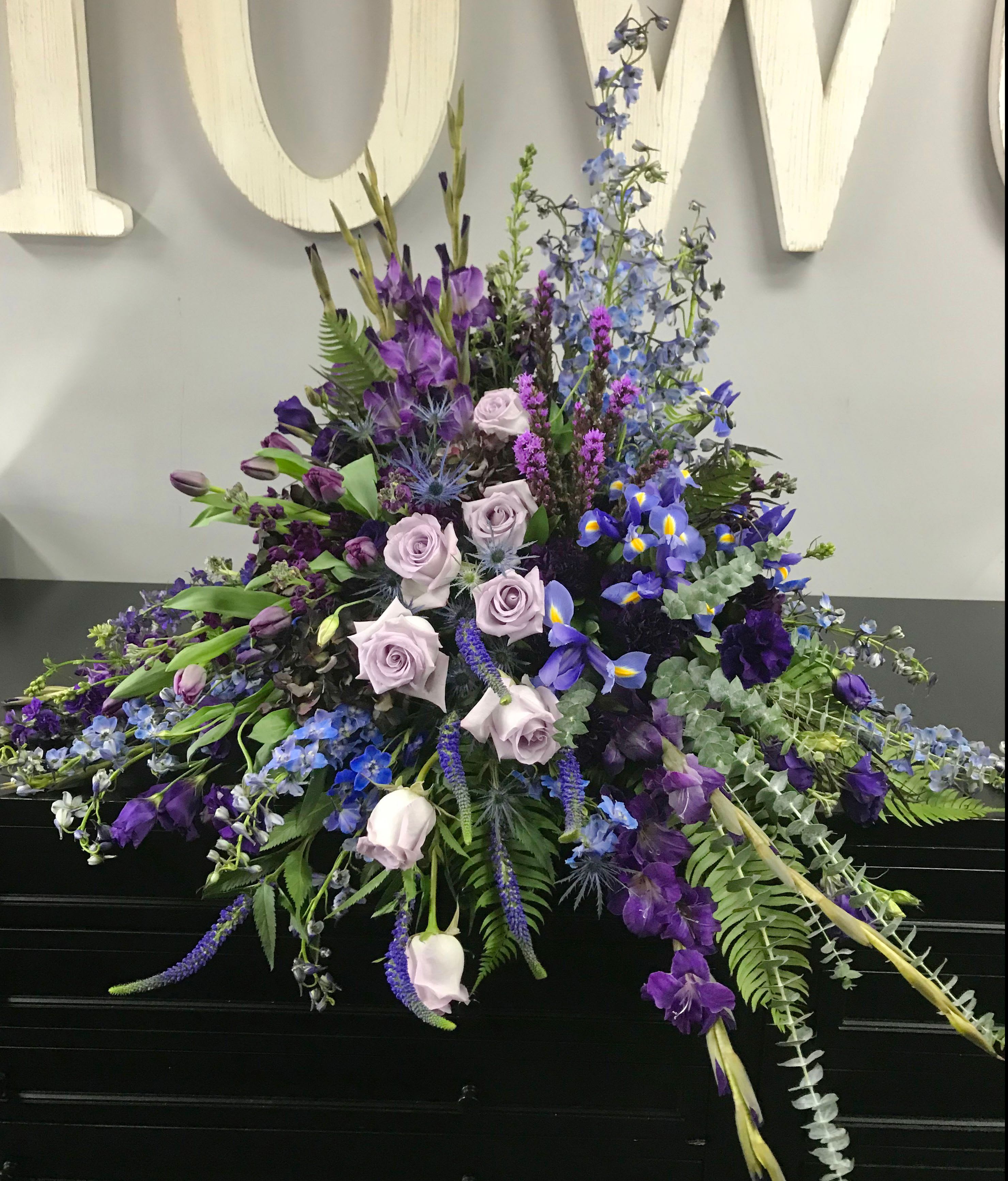 Casket Spray CS-19 - A bold and elegant expression of remembrance, this striking casket spray showcases a rich mix of purple tulips, lavender roses, blue delphinium, iris, veronica, larkspur, and thistle. Soft eucalyptus and fern accents add natural movement, while the unique layered design creates a sense of quiet depth and beauty.  Perfect for honoring a loved one with refined taste or a passion for moody, jewel-toned florals, this arrangement offers a graceful tribute filled with strength, dignity, and peace.  We welcome customizations—contact us to tailor the color palette or flower selection to best reflect your loved one’s memory.