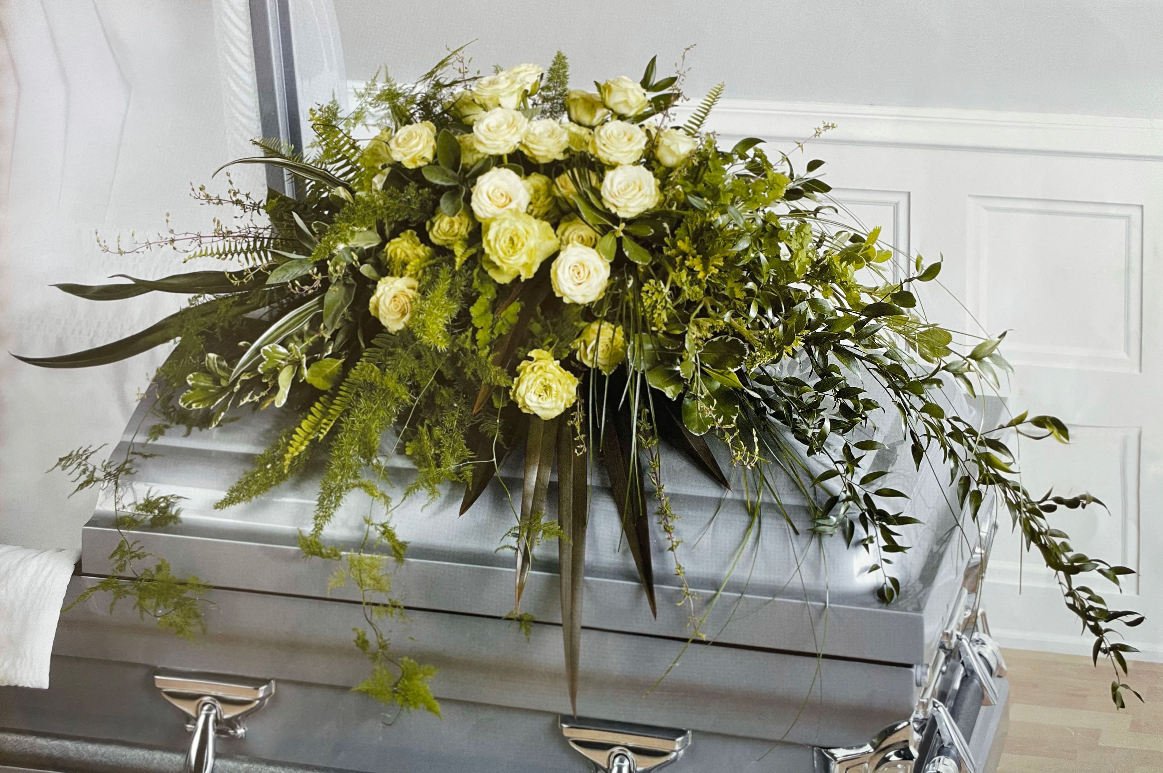 Casket Spray CS-29 - This earthy and expressive casket spray features soft cream roses surrounded by a lush array of ferns, trailing vines, and textured greenery. Designed with a fresh-from-nature look, this arrangement offers a peaceful and grounded tribute to a life lived with quiet strength and deep connection to the natural world.  Ideal for someone who appreciated simplicity, nature, or an organic style, this design brings a touch of calm beauty to the farewell.