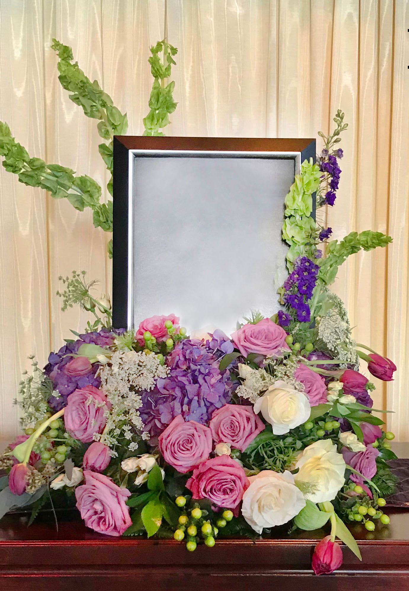 Memorial Spray MS-01 Memorial Spray MS-01 - A graceful and romantic tribute, this memorial spray features a lush arrangement of lavender roses, deep purple hydrangea, tulips, lisianthus, and creamy white blooms. Bells of Ireland and larkspur add gentle height and movement, while delicate textures like Queen Anne’s lace and hypericum provide softness and depth. Thoughtfully designed to frame a treasured photo or urn, this piece offers a heartfelt expression of love, remembrance, and peaceful reflection. (Picture frame not included.)