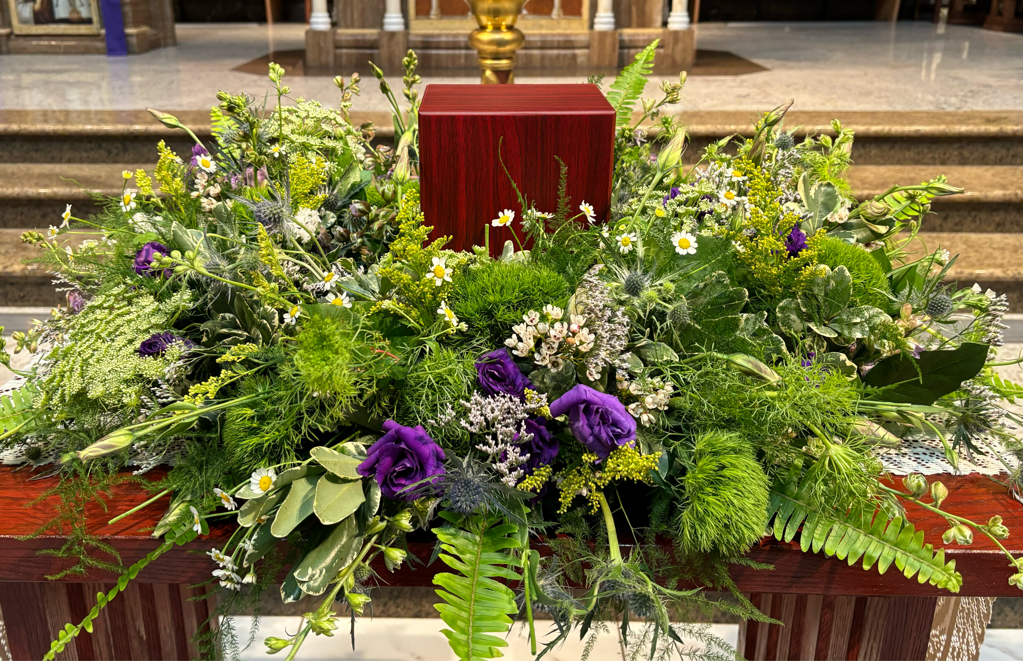 Memorial Spray MS-07 - This garden-style memorial spray evokes the gentle beauty of a peaceful meadow. A soft, flowing mix of queen anne’s lace, lisianthus, chamomile, thistle, and green dianthus is thoughtfully arranged with lush greenery and delicate filler blooms. Designed to surround an urn with a sense of serenity and natural grace, this arrangement honors a life rooted in nature, simplicity, and quiet strength. (Urn not included)