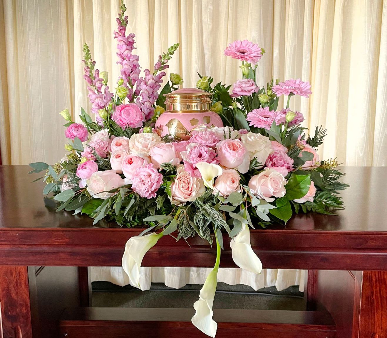 Memorial Spray MS-09 Memorial Spray MS-09 - Delicate and full of grace, this soft pink memorial spray is a tender tribute to a cherished life. Blush roses, peonies or garden roses, and ranunculus are paired with snapdragons, gerbera daisies, and creamy calla lilies, creating a romantic and serene display. Designed to cradle the urn in gentle beauty, this arrangement offers comfort and love with every bloom—perfect for honoring someone remembered with warmth and affection. (Urn not included)
