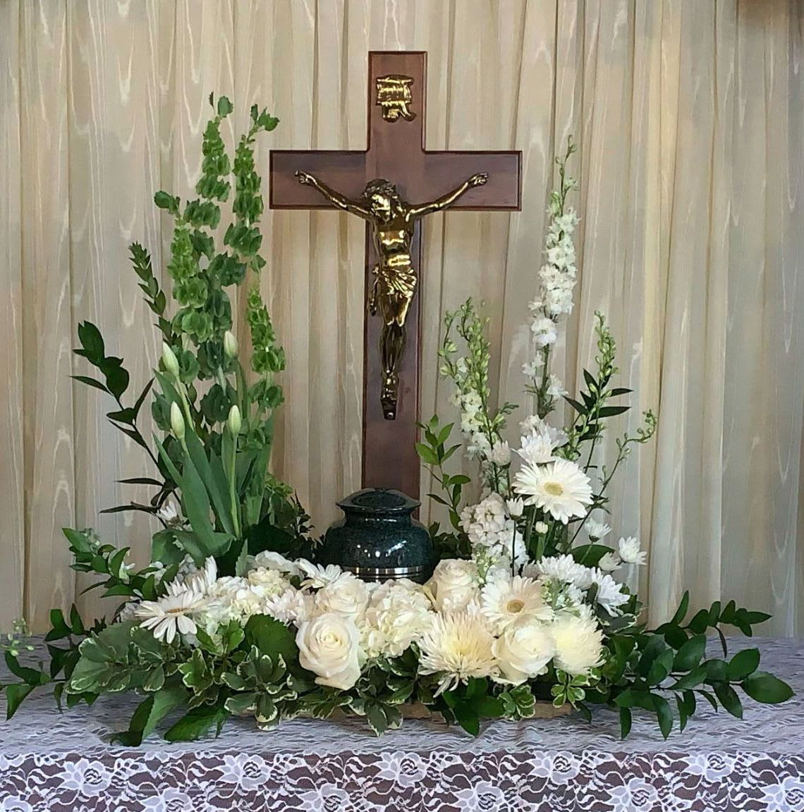 Memorial Spray MS-16 Memorial Spray MS-16 - Pure and peaceful, this all-white memorial spray offers a timeless expression of love and reverence. White roses, hydrangea, gerbera daisies, tulips, and snapdragons are artfully arranged with soft greenery and bells of Ireland for gentle height. Framing the urn, this design brings a sense of grace, faith, and tranquility—honoring a loved one with beauty and spiritual serenity. (Urn and crucifix not included)