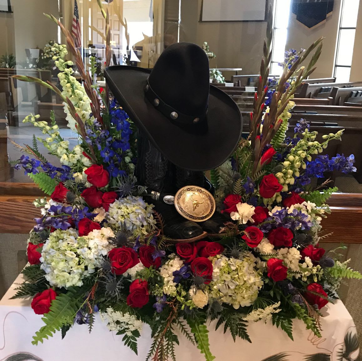 Memorial Spray MS-22 Memorial Spray MS-22 - A proud and personal tribute, this Western-themed memorial spray (boots and hat keepsakes not included) celebrates a life lived with grit, heart, and honor. Featuring classic red roses, white hydrangea, blue delphinium, thistle, and snapdragons, the design is accented with rustic greenery and natural textures. Perfect for a cowboy, rancher, or rodeo enthusiast, this arrangement beautifully frames cherished personal items while honoring a legacy rooted in strength and tradition.
