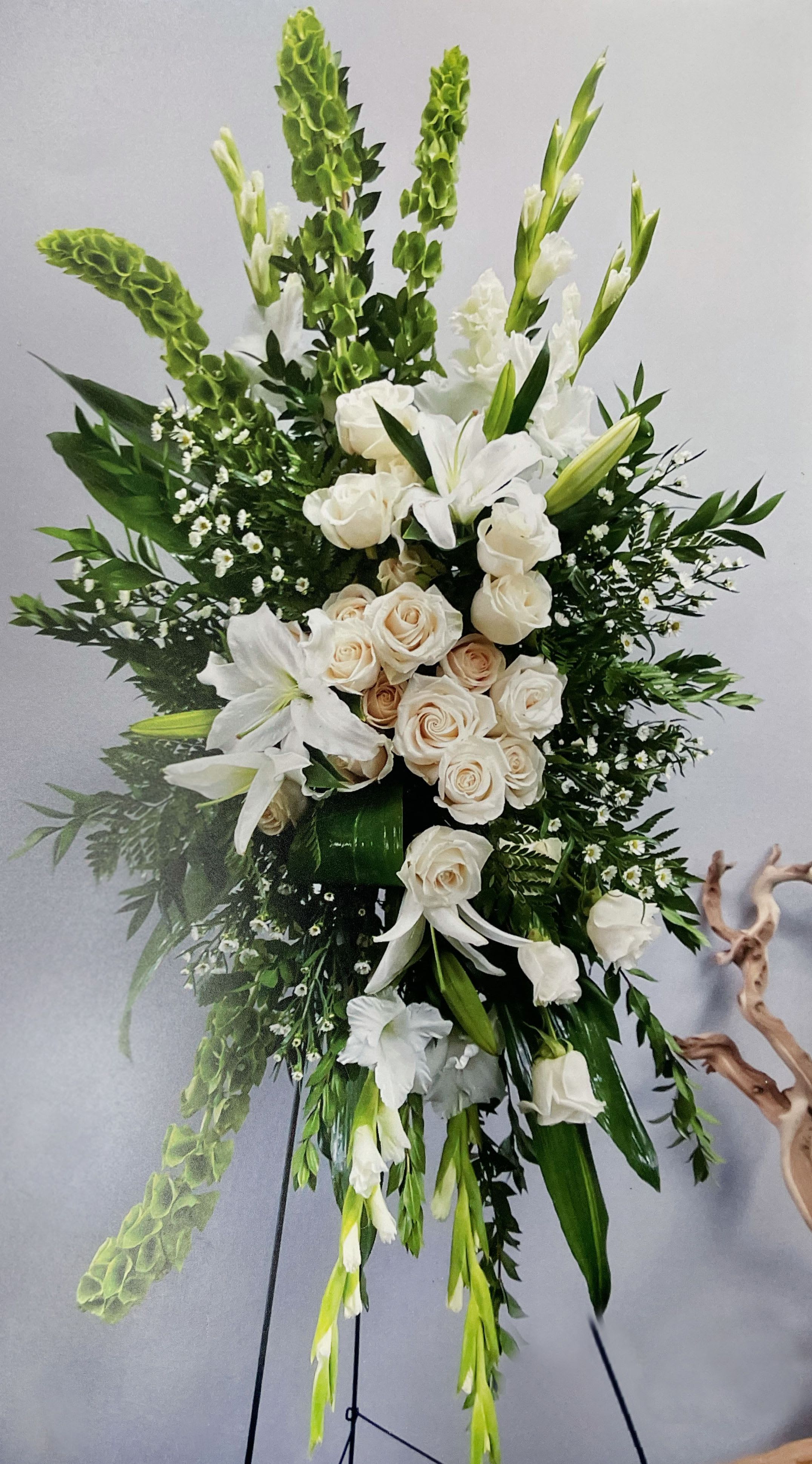 Easel Spray ES-07 Easel Spray ES-07 - Elegant and understated, this white and green easel spray offers a peaceful expression of remembrance. Featuring creamy white roses, lilies, gladiolus, and bells of Ireland, the arrangement is layered with fresh greenery and subtle textures for a graceful, vertical silhouette. Displayed on a stand (included), this design brings a touch of purity and calm to the service, perfect for honoring a loved one with quiet strength and beauty.
