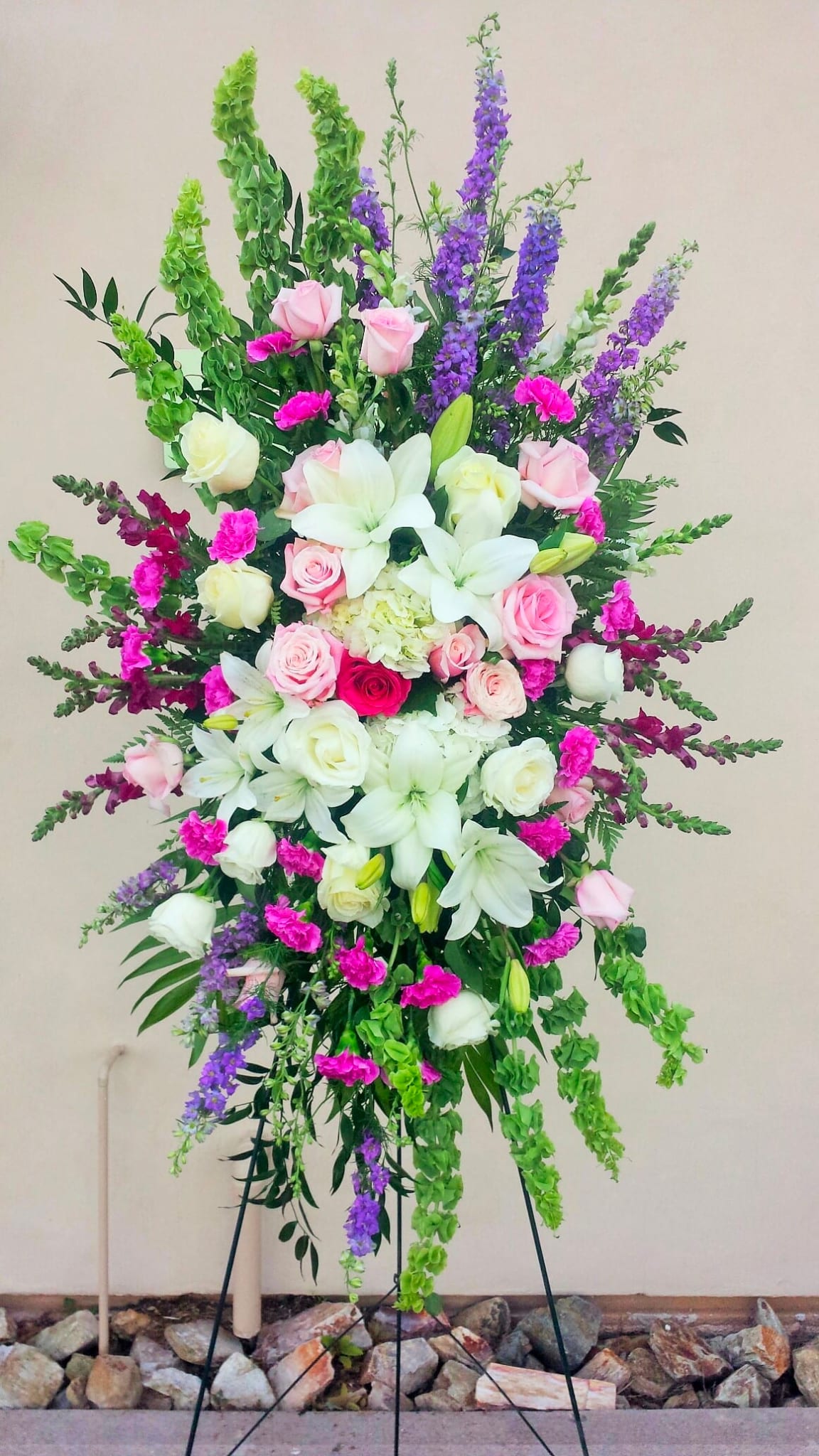 Easel Spray ES-15 - This uplifting easel spray features a soft and joyful mix of white lilies, pink roses, carnations, snapdragons, bells of Ireland, and touches of lavender and purple accents. Designed with graceful movement and garden-inspired texture, this tribute offers a message of hope, beauty, and remembrance. Presented on a standing easel (included), it brings a tender and heartfelt presence to the service.