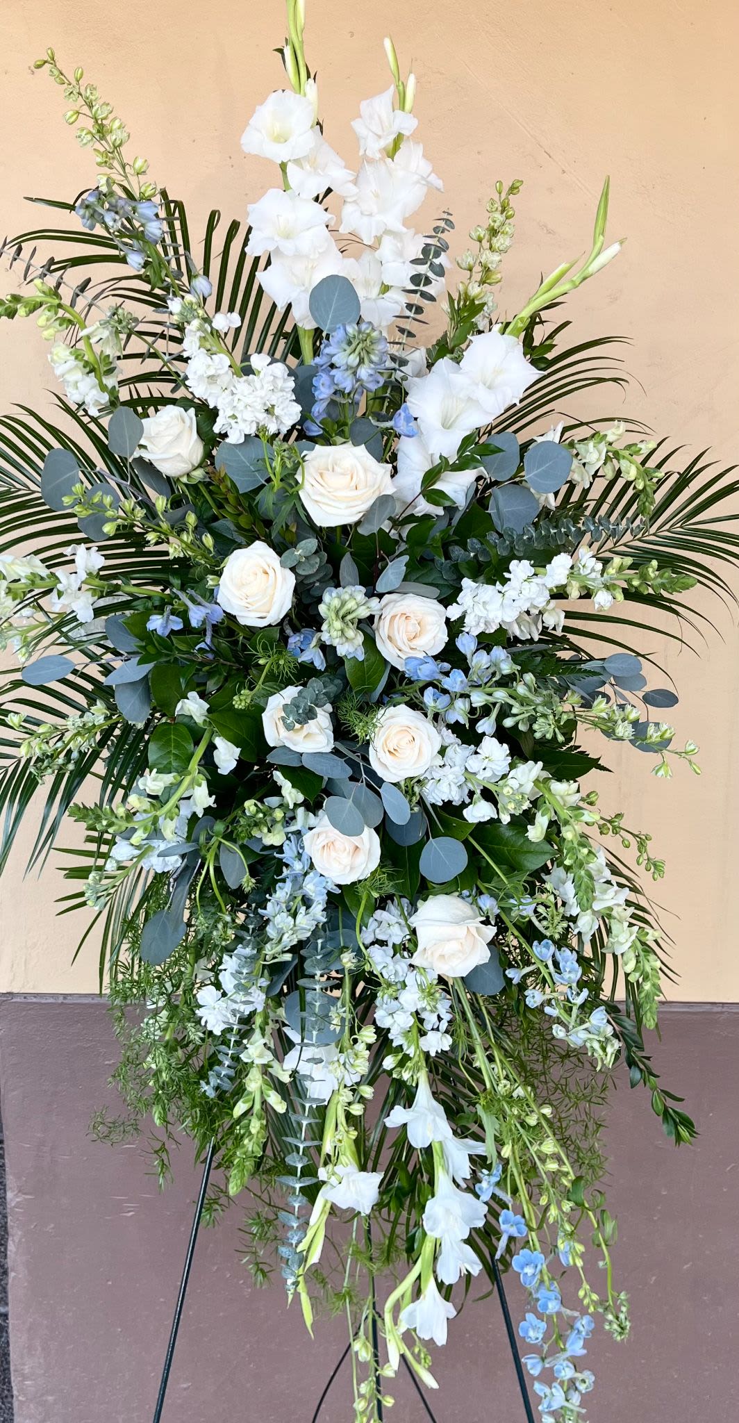 Easel Spray ES-14 - This soft and airy standing spray features a peaceful blend of ivory roses, white gladiolus, stock, snapdragons, and touches of pale blue delphinium. Accented with flowing greenery and silvery eucalyptus, it’s a gentle tribute that brings a sense of calm and remembrance. Displayed on a stand (included), this spray is a graceful expression of sympathy for a memorial, celebration of life or traditional burial funeral.