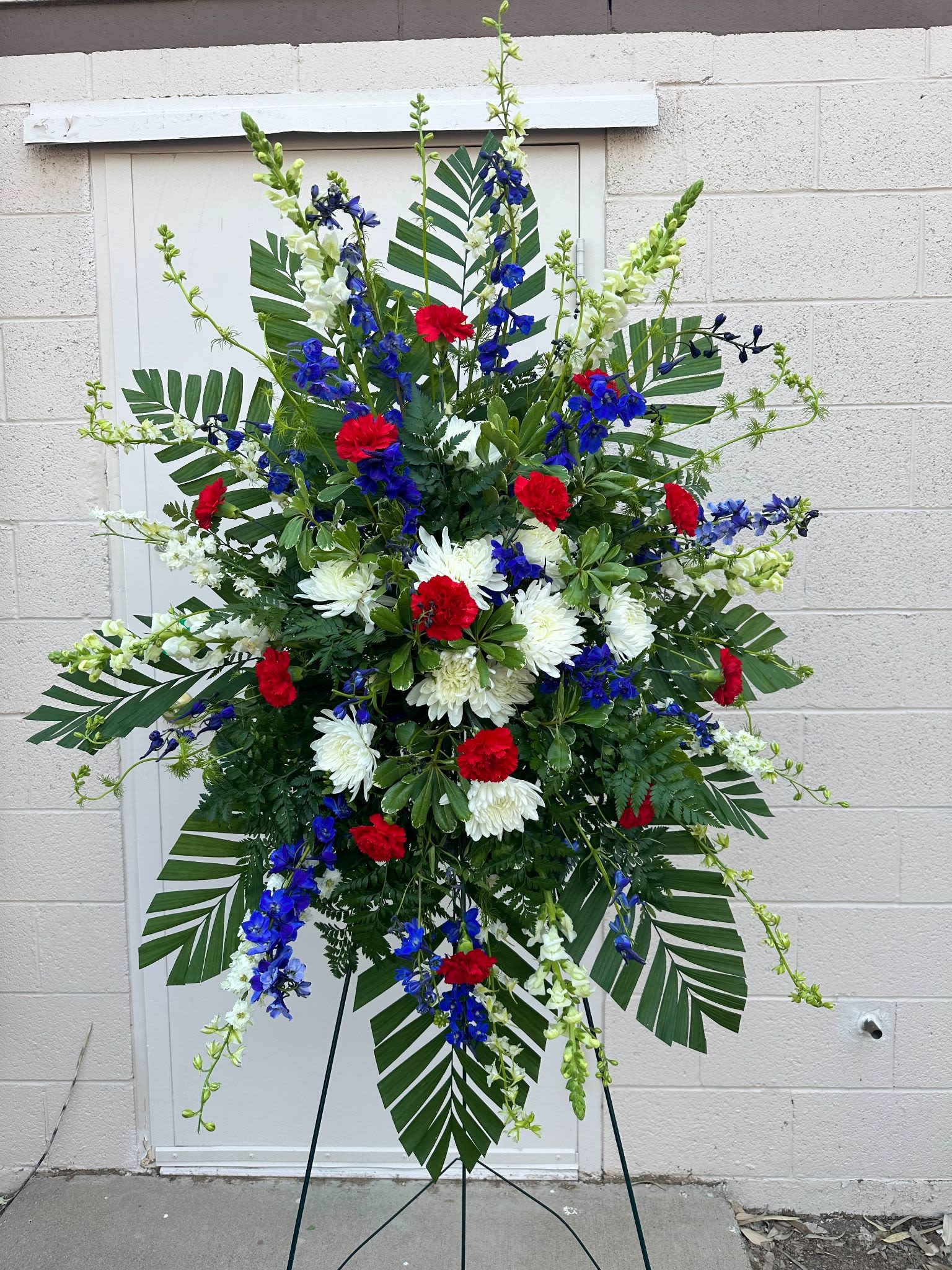 Easel Spray ES-22 Easel Spray ES-22 - This striking red, white, and blue easel spray features bold red carnations, white mums and snapdragons, and vivid blue delphinium. Accented with tropical palm leaves and lush greenery, it's a fitting tribute for honoring military service, patriotic spirit, or a life well lived. Easel stand included.