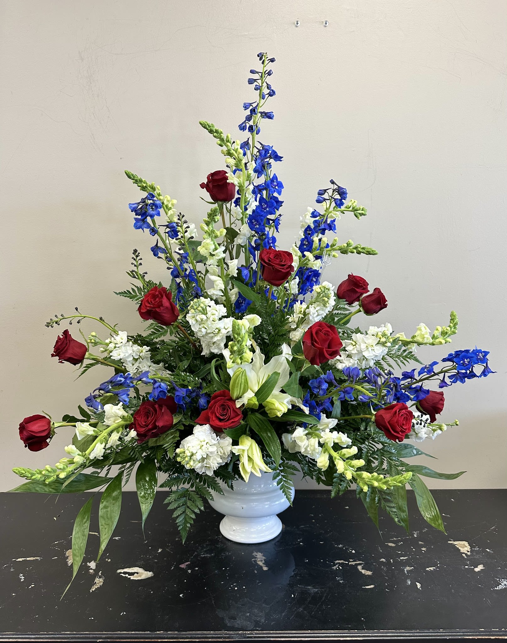 Ceremony Arrangement CV-15 Ceremony Arrangement CV-15 - A bold and heartfelt tribute featuring classic red roses, white lilies and snapdragons, and vivid blue delphinium. This ceremony arrangement reflects strength, dignity, and patriotic pride—perfect for honoring a veteran or loved one with deep American roots.